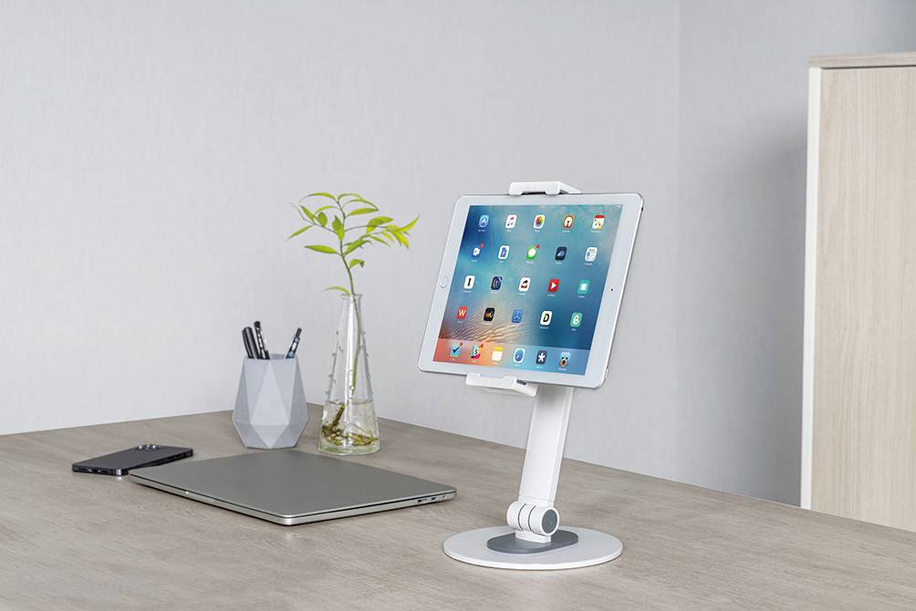Auf einem Schreibtisch steht ein Tablet in einem weißen Ständer neben einem Laptop, einem Smartphone und einer Vase mit Zweigen.