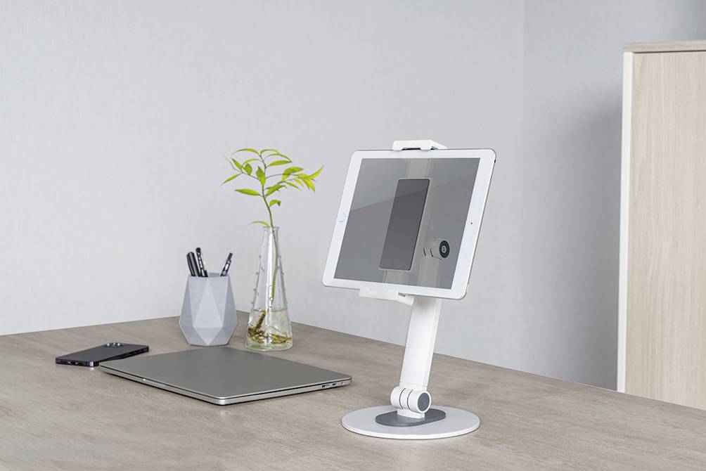 Ein Tablet steht auf einem weißen Ständer auf einem Schreibtisch. Daneben liegen ein Laptop und ein Smartphone. Im Hintergrund eine Vase mit Pflanze.