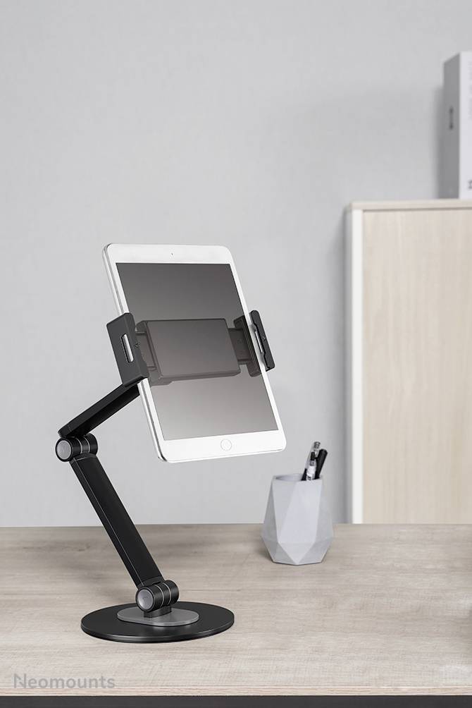 Ein Tablet steht in einem verstellbaren schwarzen Halter mit Arm auf einem Schreibtisch. Im Hintergrund sind ein Stifthalter und ein Schrank.