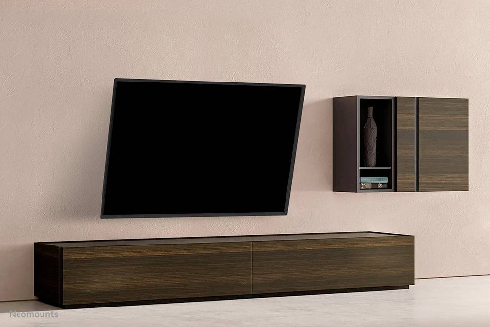 Ein moderner Fernseher hängt schräg an einer rosa Wand. Darunter steht ein flaches, dunkles Holzregal. Rechts oben ein kleines Wandregal.