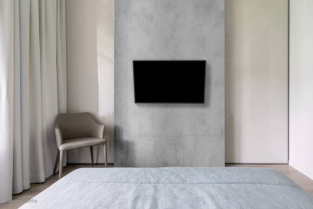 Ein Schlafzimmer mit einem grauen Bett, einem Fernseher an der Wand und einem braunen Stuhl in der Ecke neben einem Fenster mit Vorhängen.