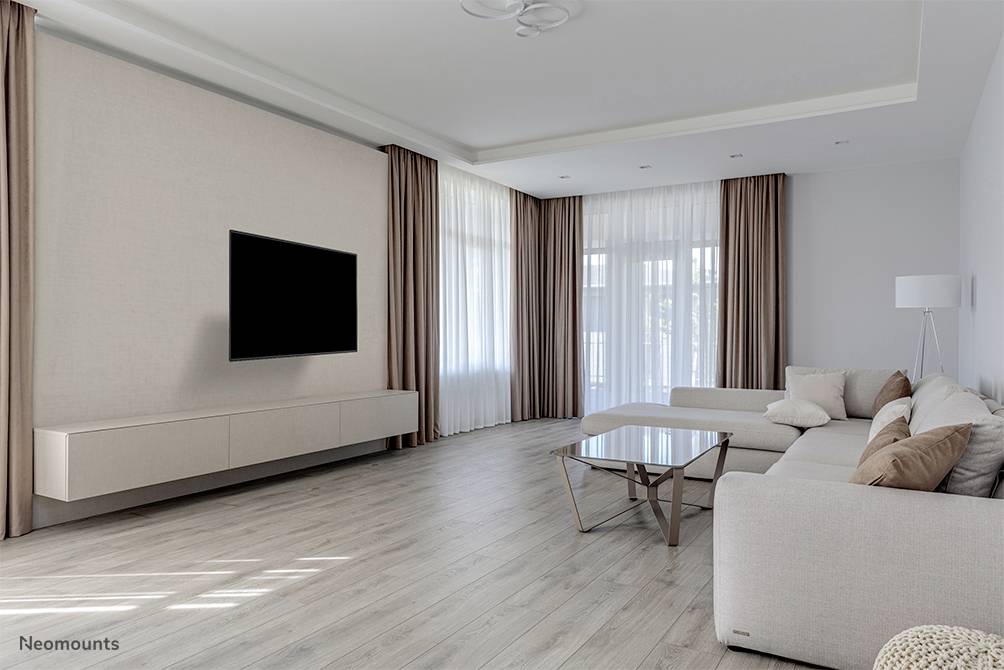 Helles Wohnzimmer mit modernem Sofa, Couchtisch und großem Wandfernseher, gerahmt von bodenlangen Vorhängen vor großen Fenstern.