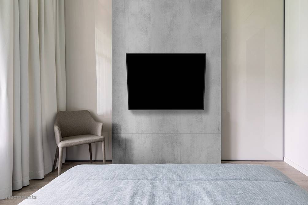 Ein modernes Schlafzimmer mit grauer Wand, an der ein großer Flachbildfernseher hängt. Ein Stuhl steht in der Ecke neben hellen Vorhängen.