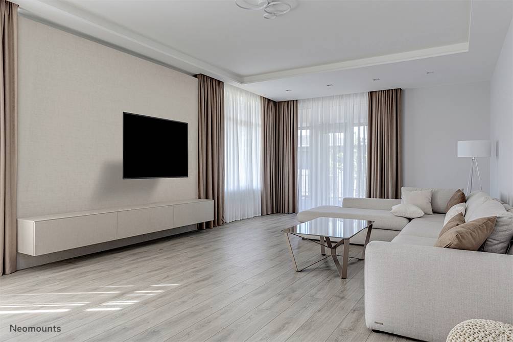 Helles Wohnzimmer mit großem Ecksofa, Glastisch und Wand-TV. Große Fenster mit bodenlangen Vorhängen lassen viel Licht herein.