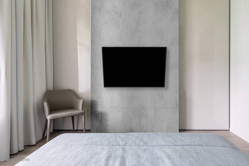 Ein modernes Schlafzimmer mit einem grauen Betonakzent an der Wand, auf dem ein großer, schwarzer Flachbildfernseher montiert ist. Ein Stuhl steht links.