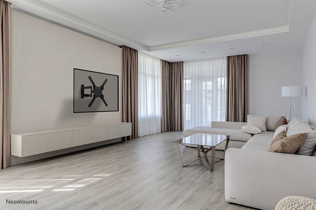 Wohnzimmer mit hellen Wänden und Holzboden. Ein großes Sofa mit Kissen steht vor einem TV-Wandhalter ohne Fernseher. Große Fenster mit langen Vorhängen lassen Licht herein.