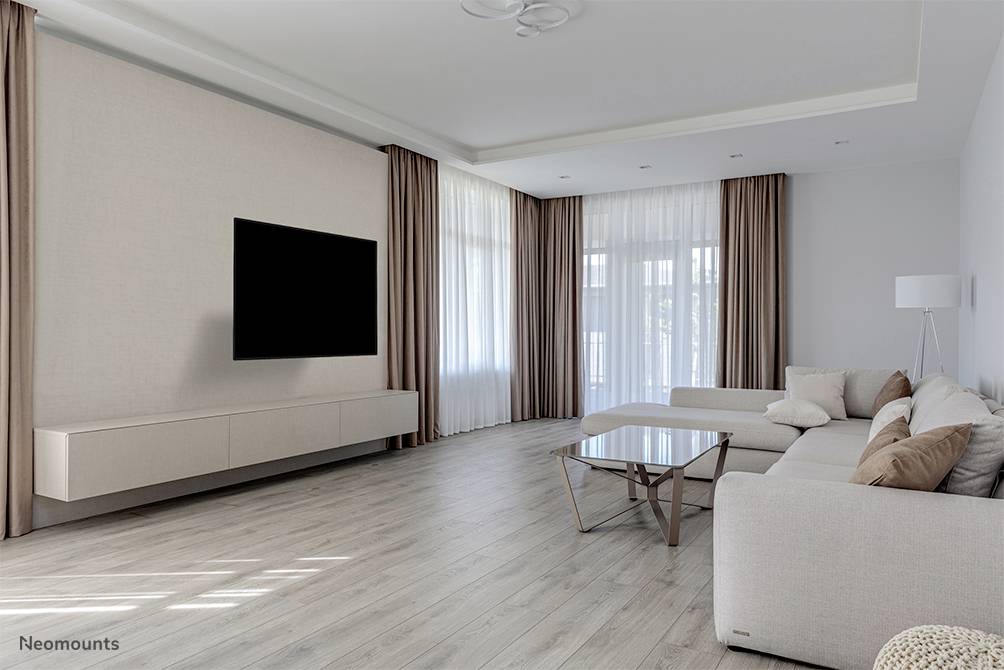 Großes Wohnzimmer mit hellem Holzboden, beiger Couch mit Kissen, Glastisch, einem an der Wand montierten Fernseher und großen Fenstern.