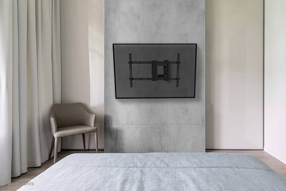 Schlafzimmer mit grauer Wand, Fernseher an der Wandhalterung, ein Stuhl links neben einem Fenster, Bett im Vordergrund.