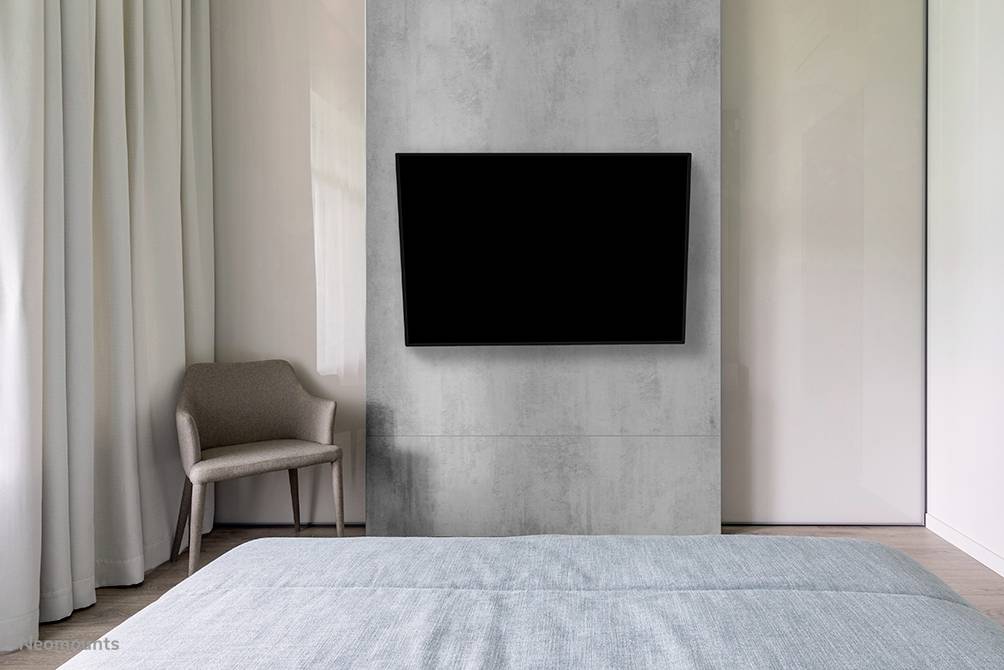 Ein modernes Schlafzimmer mit einem Fernseher an der Wand, einem Stuhl und einem Bett im Vordergrund. Neutrale Farben dominieren den Raum.