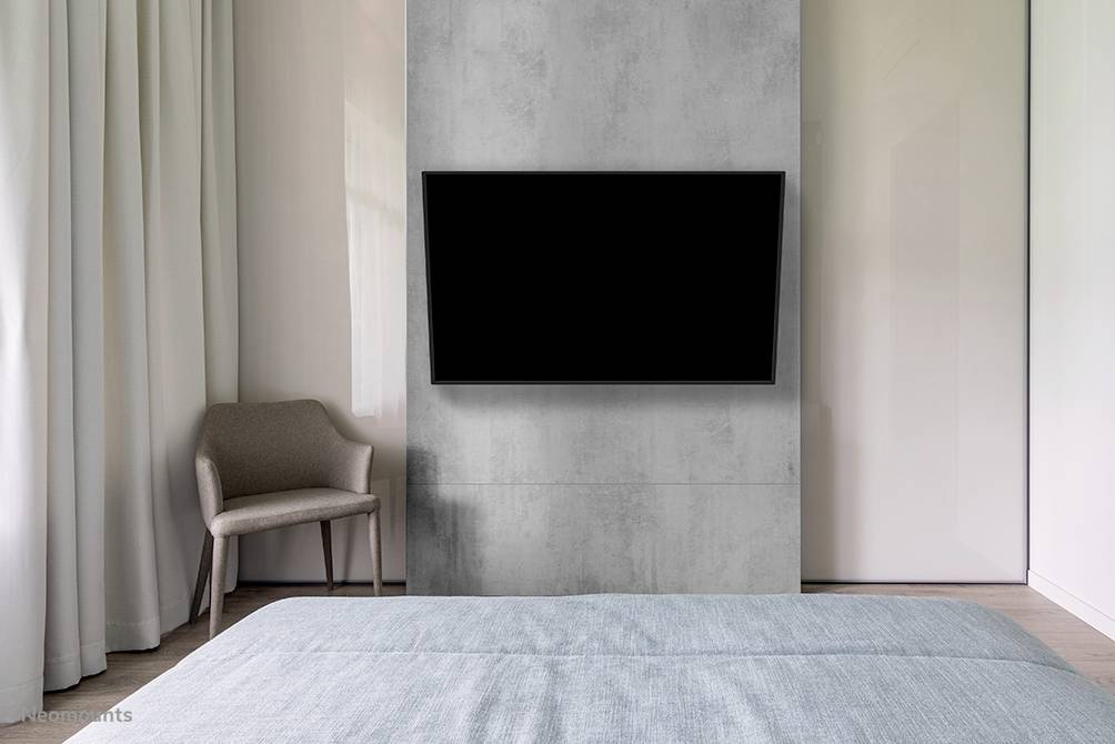Schlichtes Schlafzimmer mit grauer Wand, auf der ein Flachbildfernseher montiert ist. Links steht ein Sessel neben langen Vorhängen.