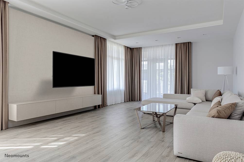 Helles Wohnzimmer mit großem Sofa, Couchtisch und wandmontiertem Fernseher. Große Fenster mit Vorhängen und Holzboden.