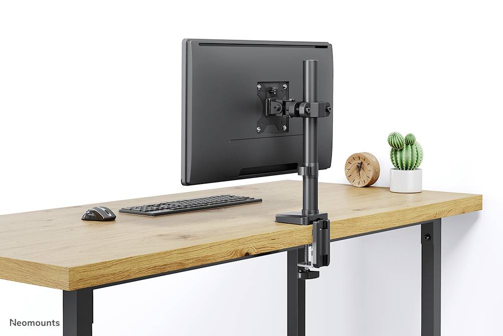 Monitorhalterung auf einem Holztisch mit Tastatur und Maus. Im Hintergrund eine Pflanze und eine Uhr.