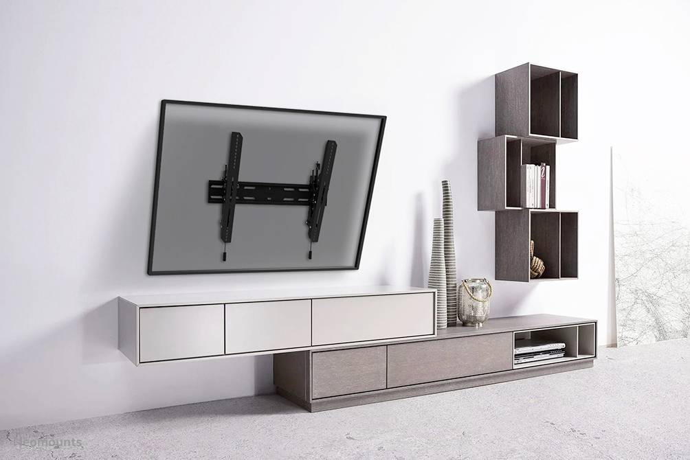 Ein Wohnzimmer mit heller Wand, an der ein schräger Fernseher auf einer Halterung montiert ist. Daneben ein modernes Regal mit Dekoration.