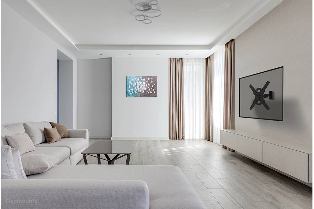 Helles Wohnzimmer mit grauem Sofa, Couchtisch, Wanddekoration und TV-Halterung an der Wand. Große Fenster mit beigen Vorhängen.