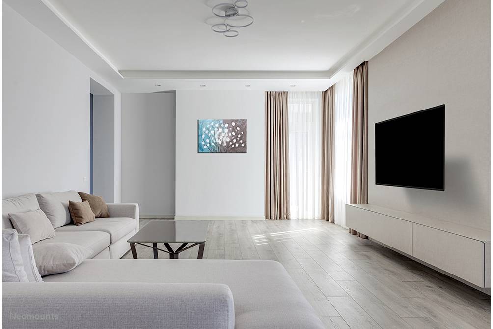 Helles Wohnzimmer mit grauem Sofa, modernem Couchtisch und großem Flachbildfernseher an der Wand, rechts große Fenster mit Vorhängen.