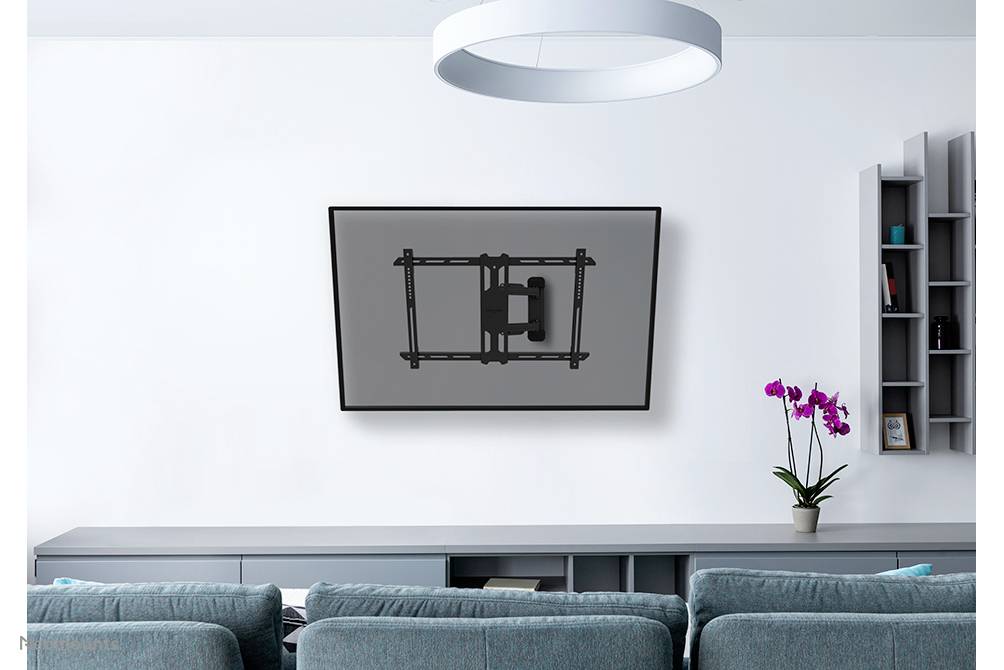 Ein moderner Wohnraum mit grauem Sofa im Vordergrund. An der weißen Wand hängt ein montierter Fernseher, daneben ein Regal mit einer Orchidee.