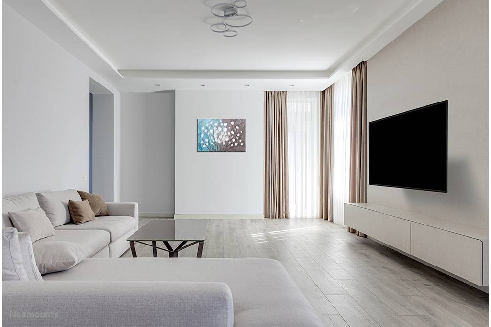 Helles Wohnzimmer mit beigem Sofa, Couchtisch, großem Fernseher an der Wand und abstraktem Kunstwerk. Große Fenster mit Vorhängen.