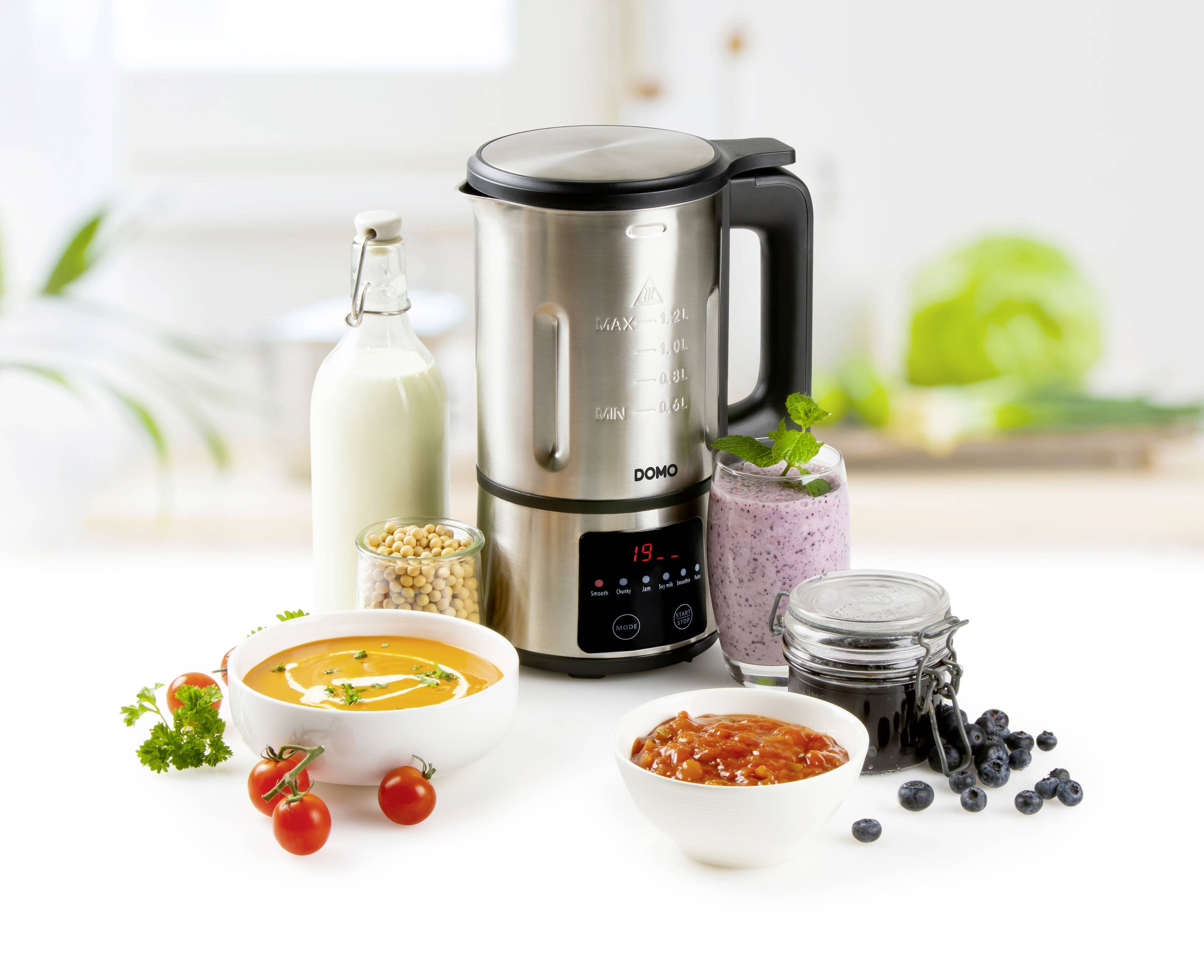 Ein Mixer steht auf einem Tisch, umgeben von einer Flasche Milch, einer Schüssel Suppe, einer Schüssel Sauce, einem Glas mit Beeren und Kichererbsen.