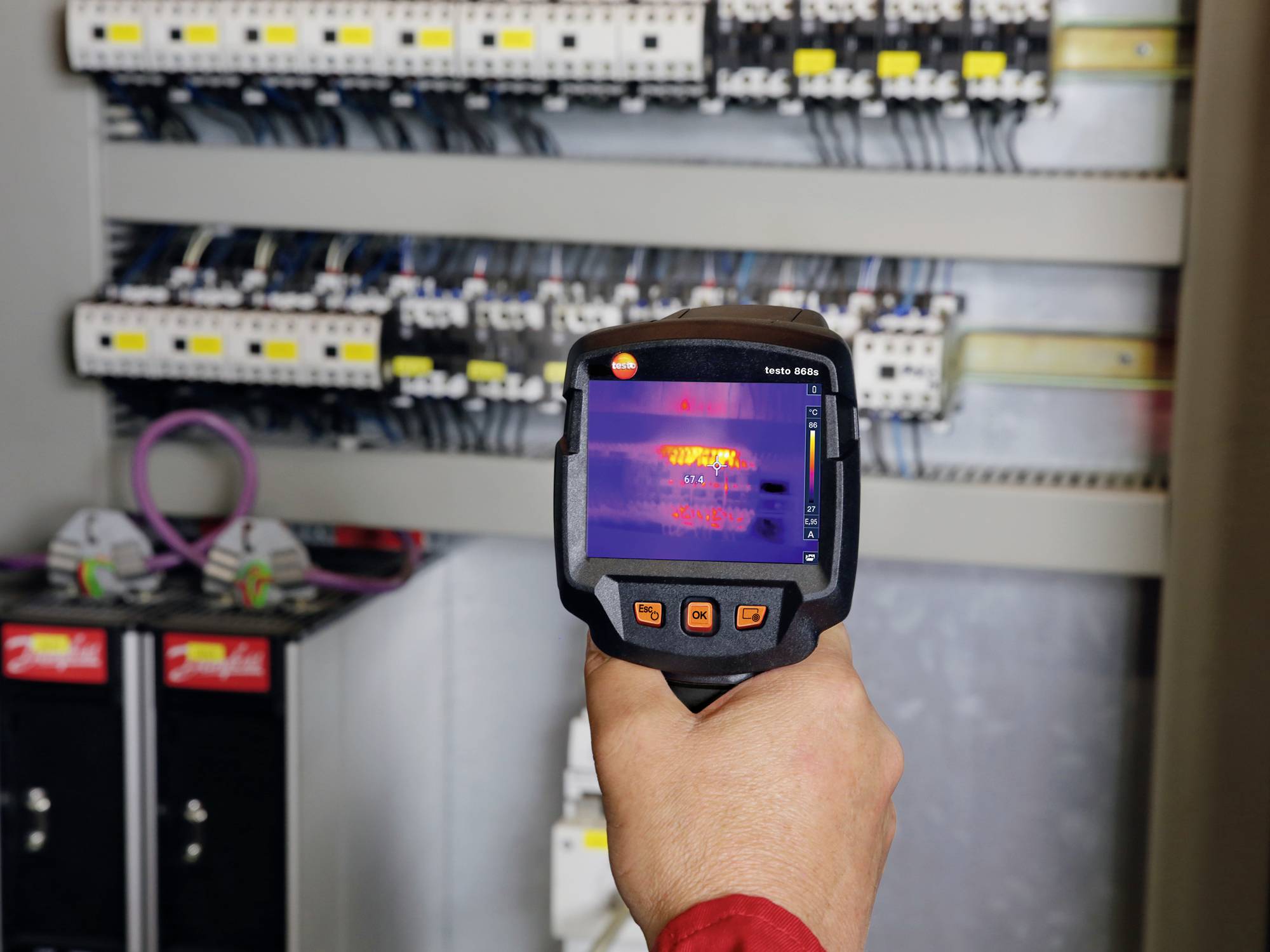 Eine Person hält eine Wärmebildkamera vor einem offenen Schaltschrank. Der Bildschirm zeigt Temperaturunterschiede an den Komponenten.