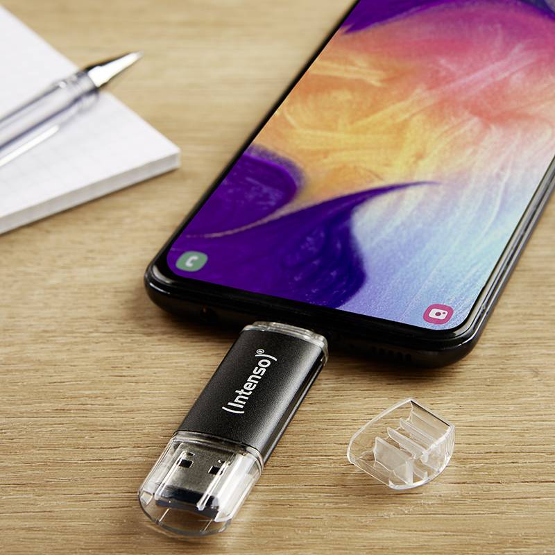 Smartphone liegt auf Holztisch, verbunden mit USB-Stick. Neben ihm liegt ein Notizblock mit einem Stift. Bildschirm zeigt bunte Muster.