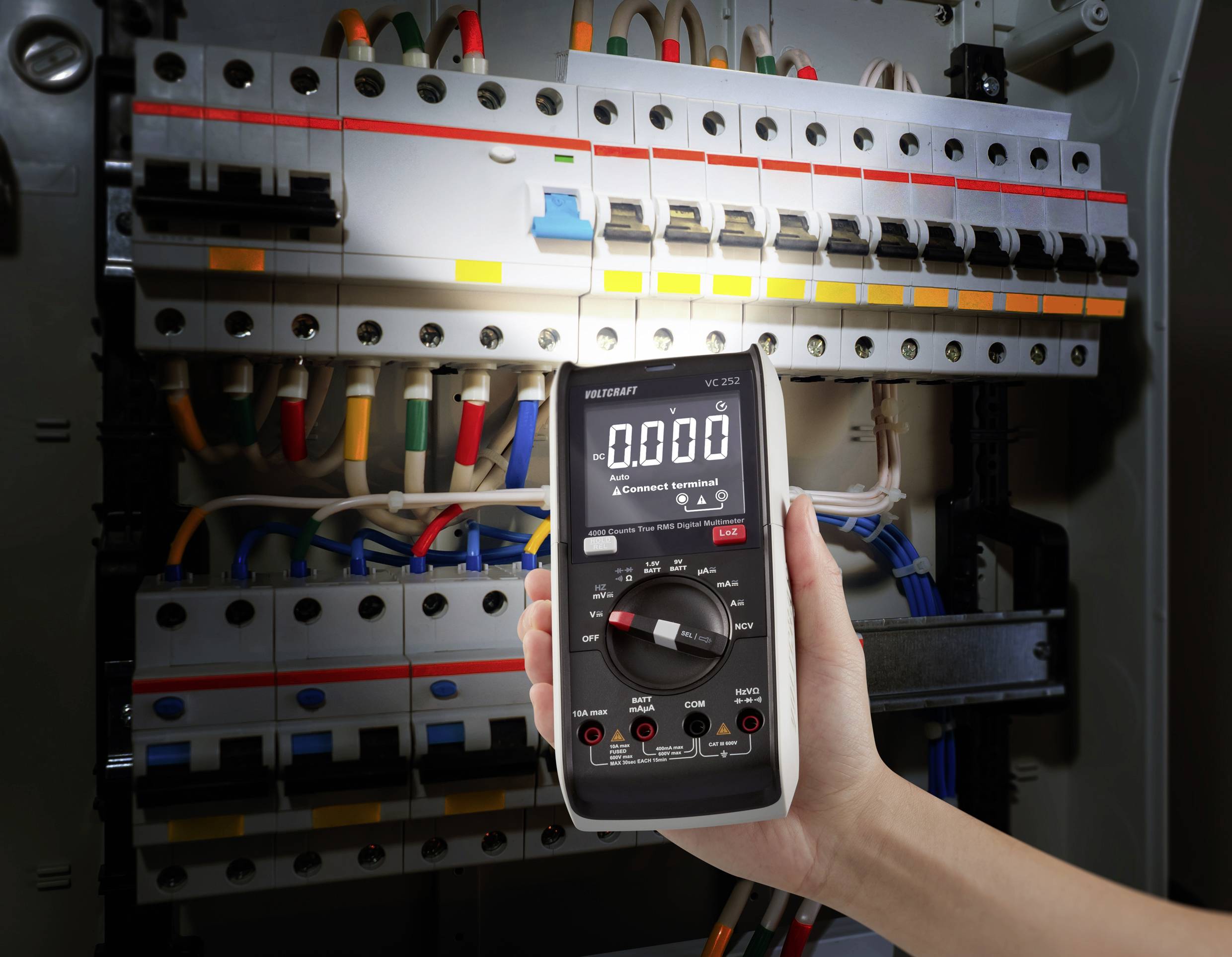 Eine Hand hält ein digitales Multimeter, das eine Spannung von 0,000 anzeigt, vor einem geöffneten Stromverteilerkasten mit mehreren Kabeln.