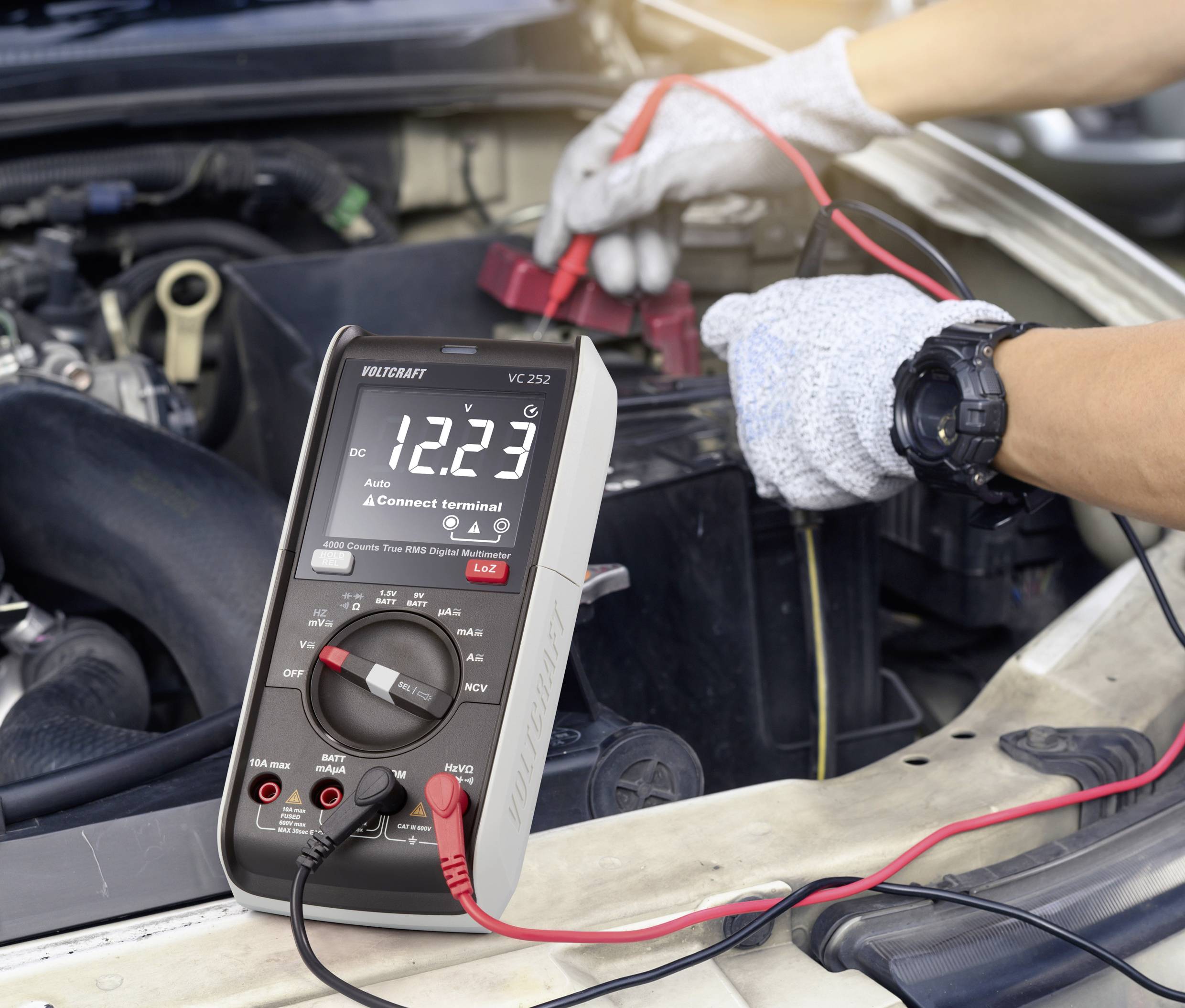 Ein Multimeter zeigt 12,23 Volt an, während es an eine Autobatterie im Motorraum angeschlossen ist. Eine Person in Handschuhen hält die Klemmen.