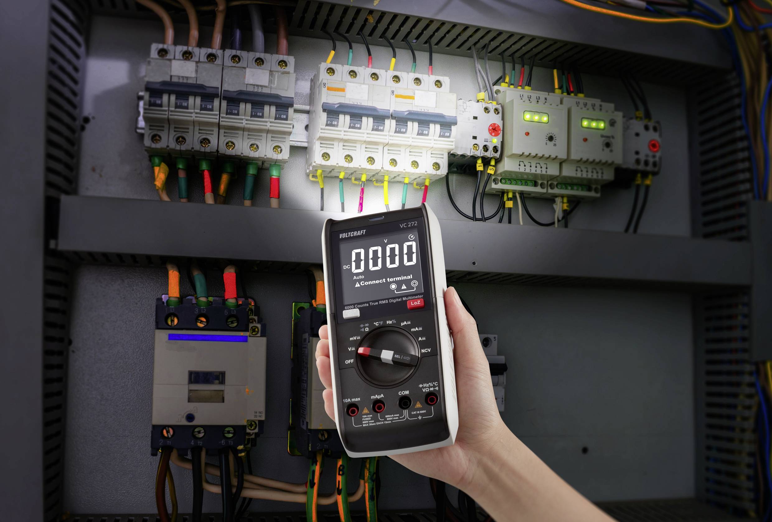 Eine Hand hält ein digitales Multimeter, das auf Null anzeigt, vor einem geöffneten elektrischen Schaltschrank mit Sicherungen und Kabeln.