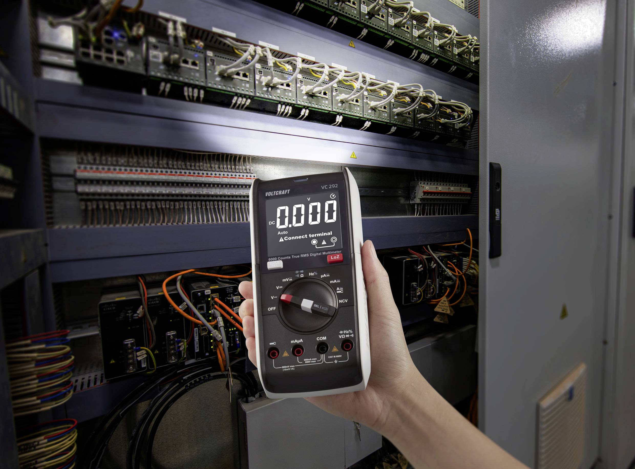 Eine Hand hält ein Digitalmultimeter, das auf 0.000 eingestellt ist, vor einem offenen Schaltschrank voller Kabel und elektronischer Bauteile.