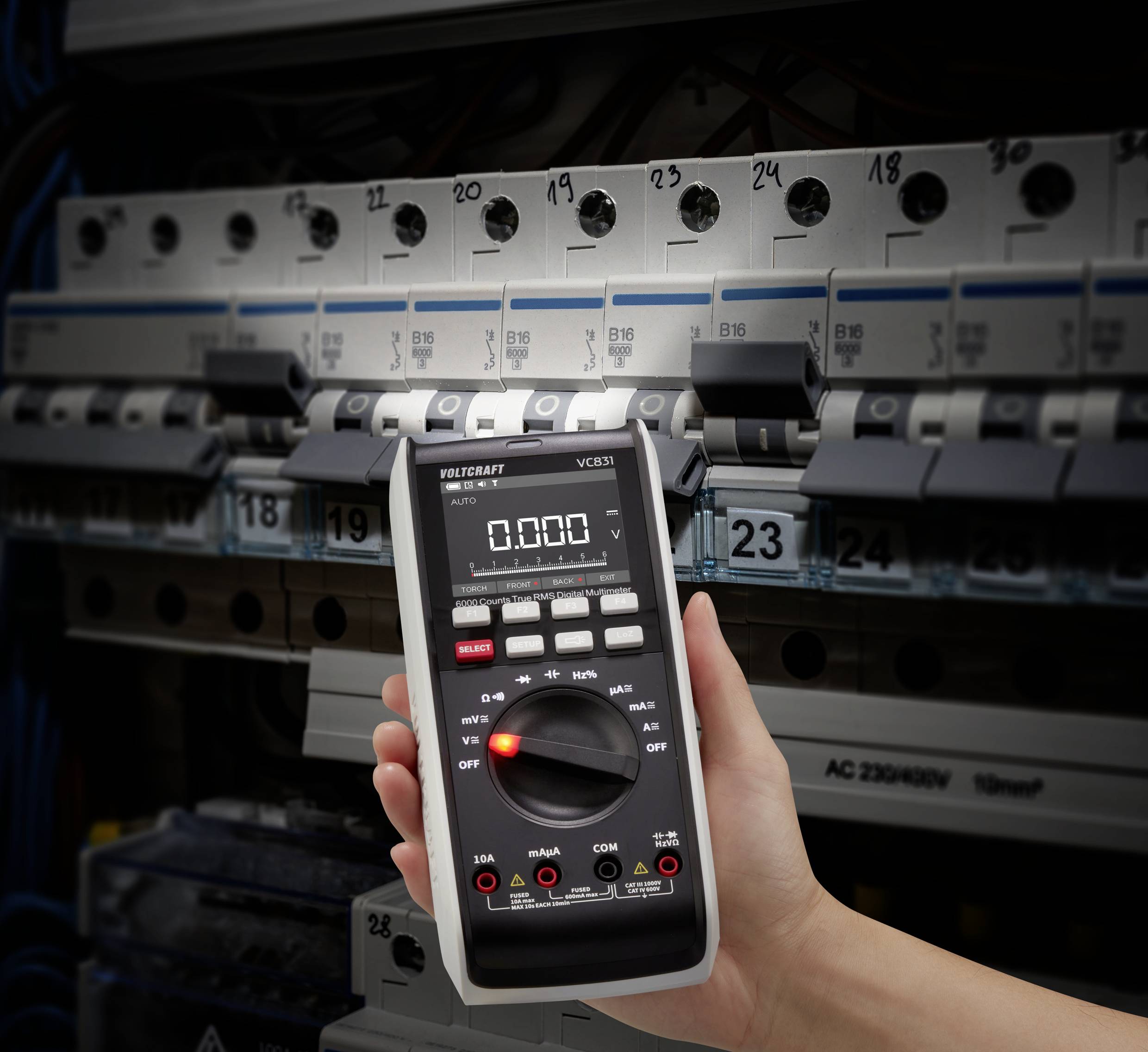 Eine Hand hält ein digitales Multimeter vor einem geöffneten Sicherungskasten mit mehreren Sicherungsautomaten.