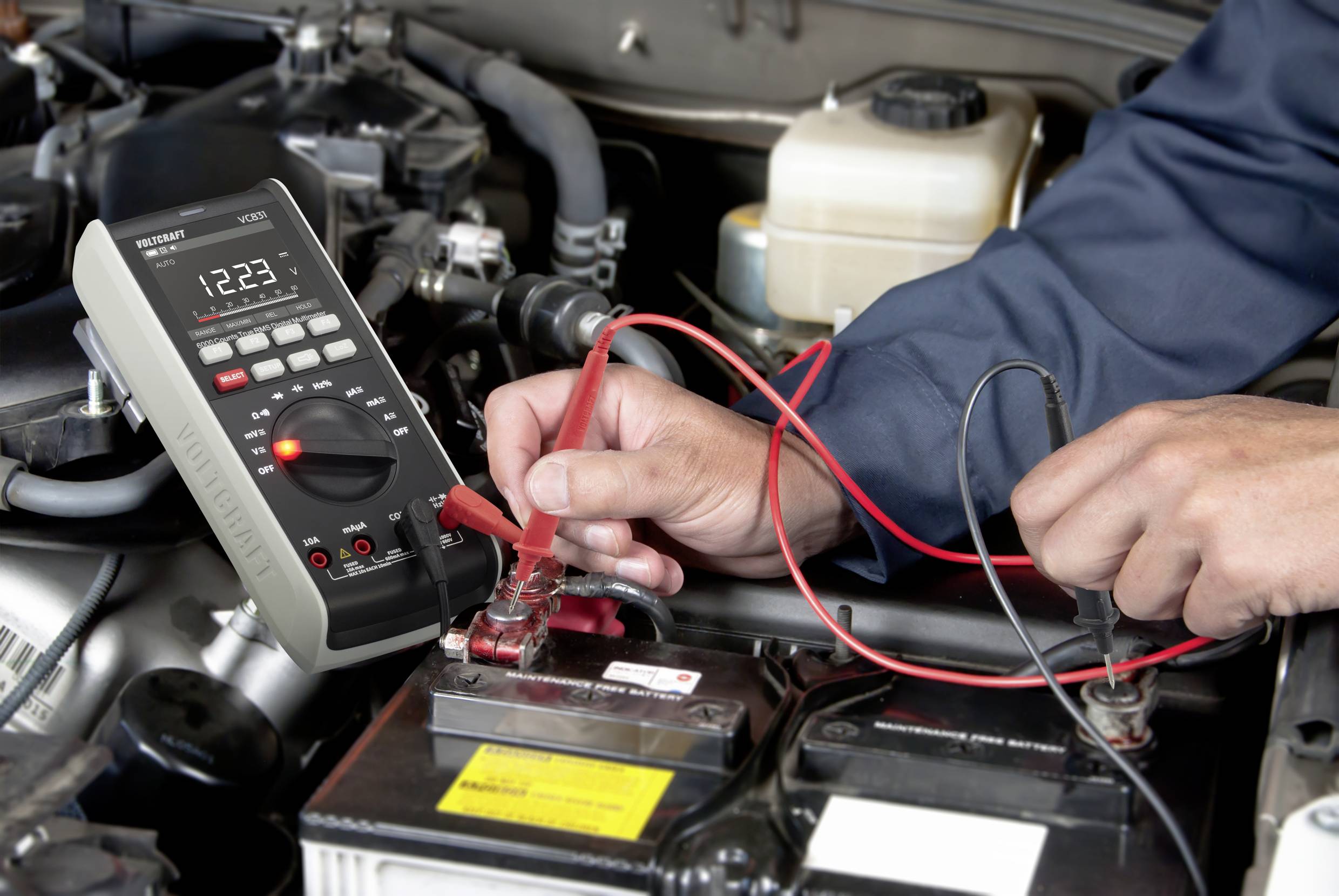 Eine Person misst mit einem digitalen Multimeter die Spannung einer Autobatterie im Motorraum. Fokus auf die Hände und das Messgerät.