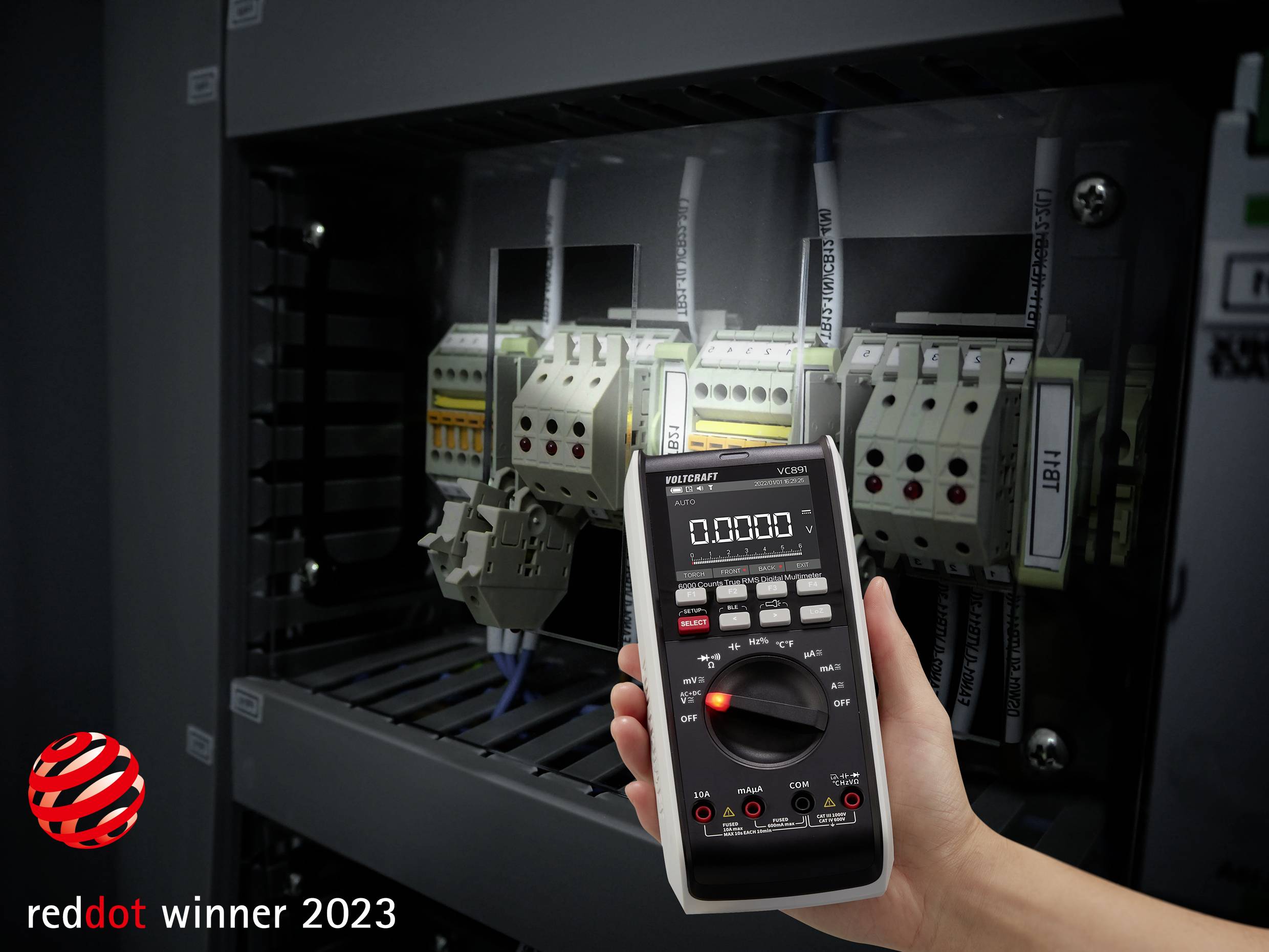 Eine Hand hält ein digitales Multimeter vor einem Schaltschrank mit elektrischen Bauteilen, 'reddot winner 2023' in der Ecke.