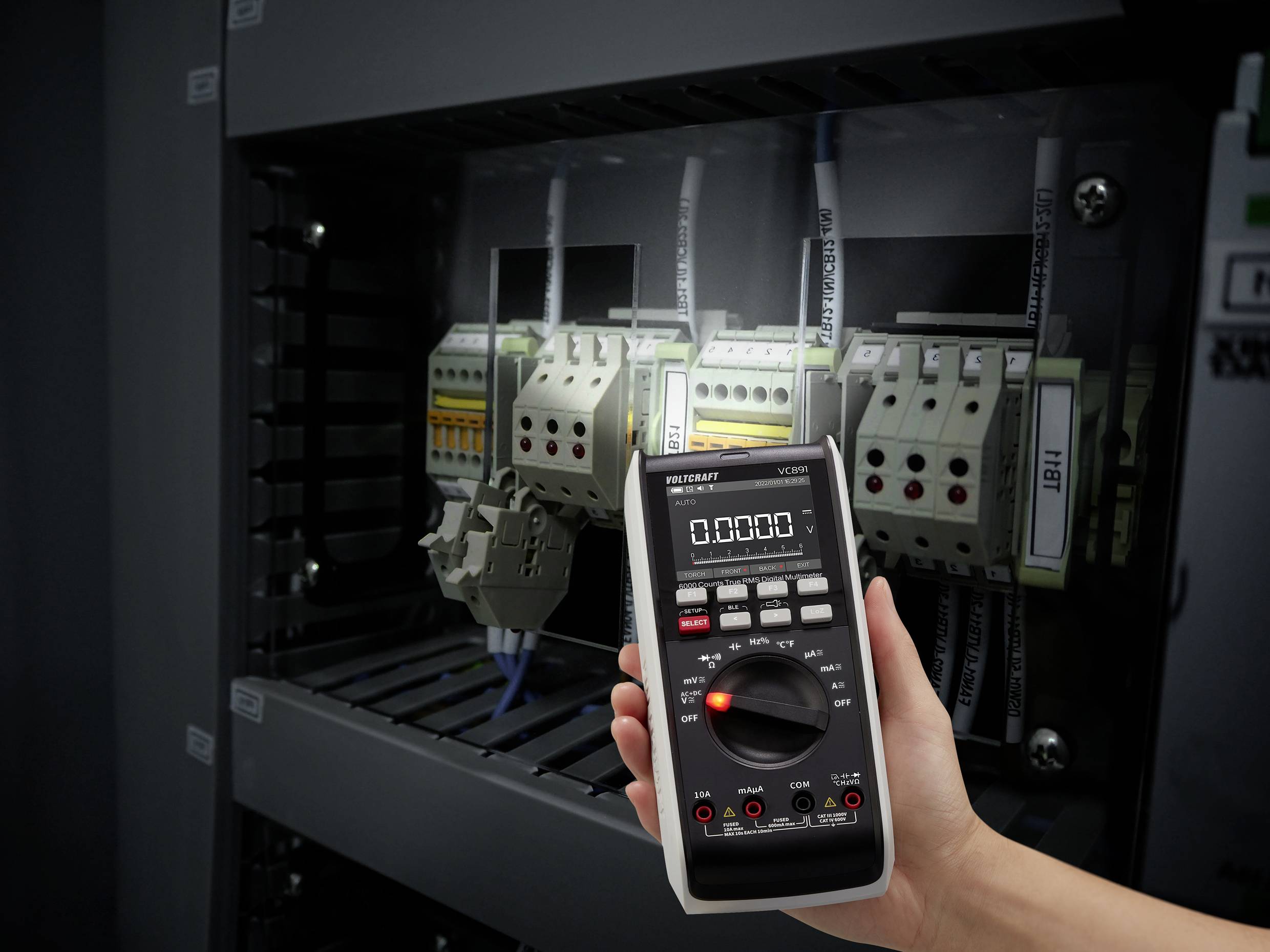 Eine Hand hält ein Multimeter vor einem Schaltschrank, in dem mehrere elektrische Bauteile und Kabel sichtbar sind.