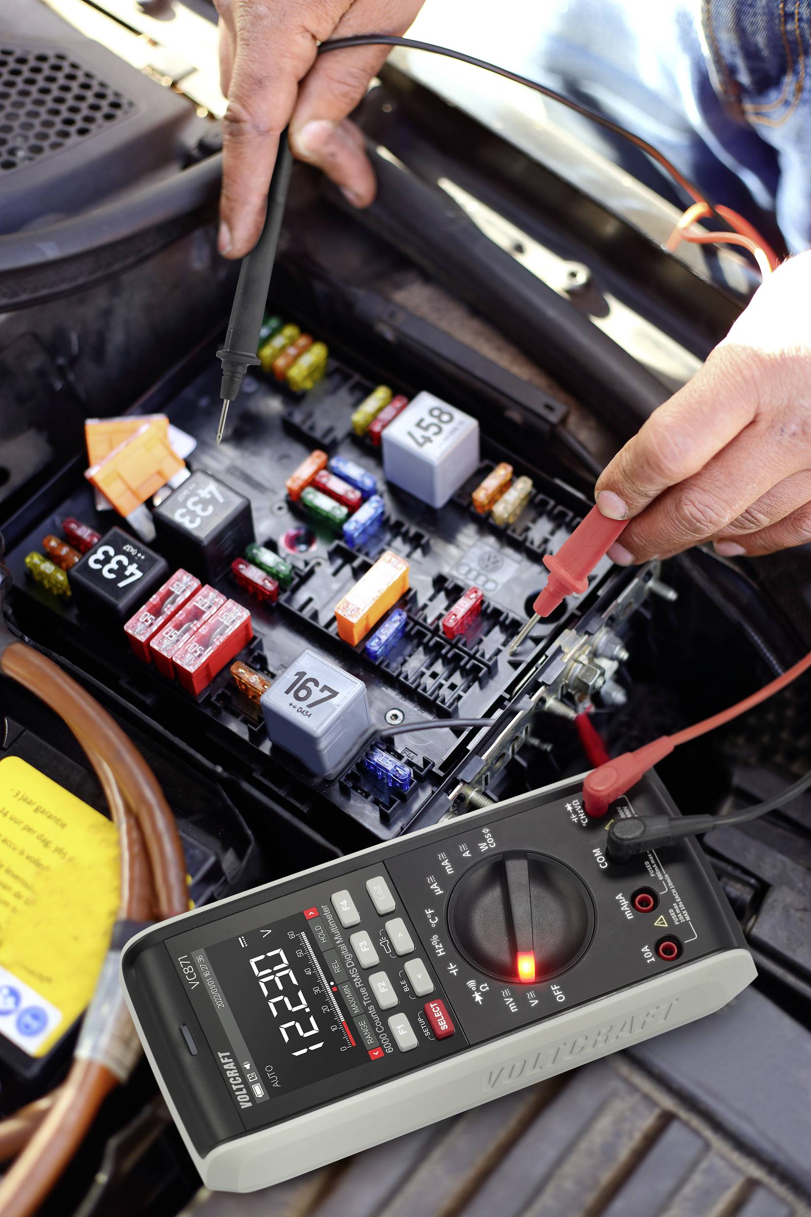 Eine Person misst mit einem Multimeter die Spannung in einer Autosicherungskasten. Der Messwert zeigt 12,27 Volt an.