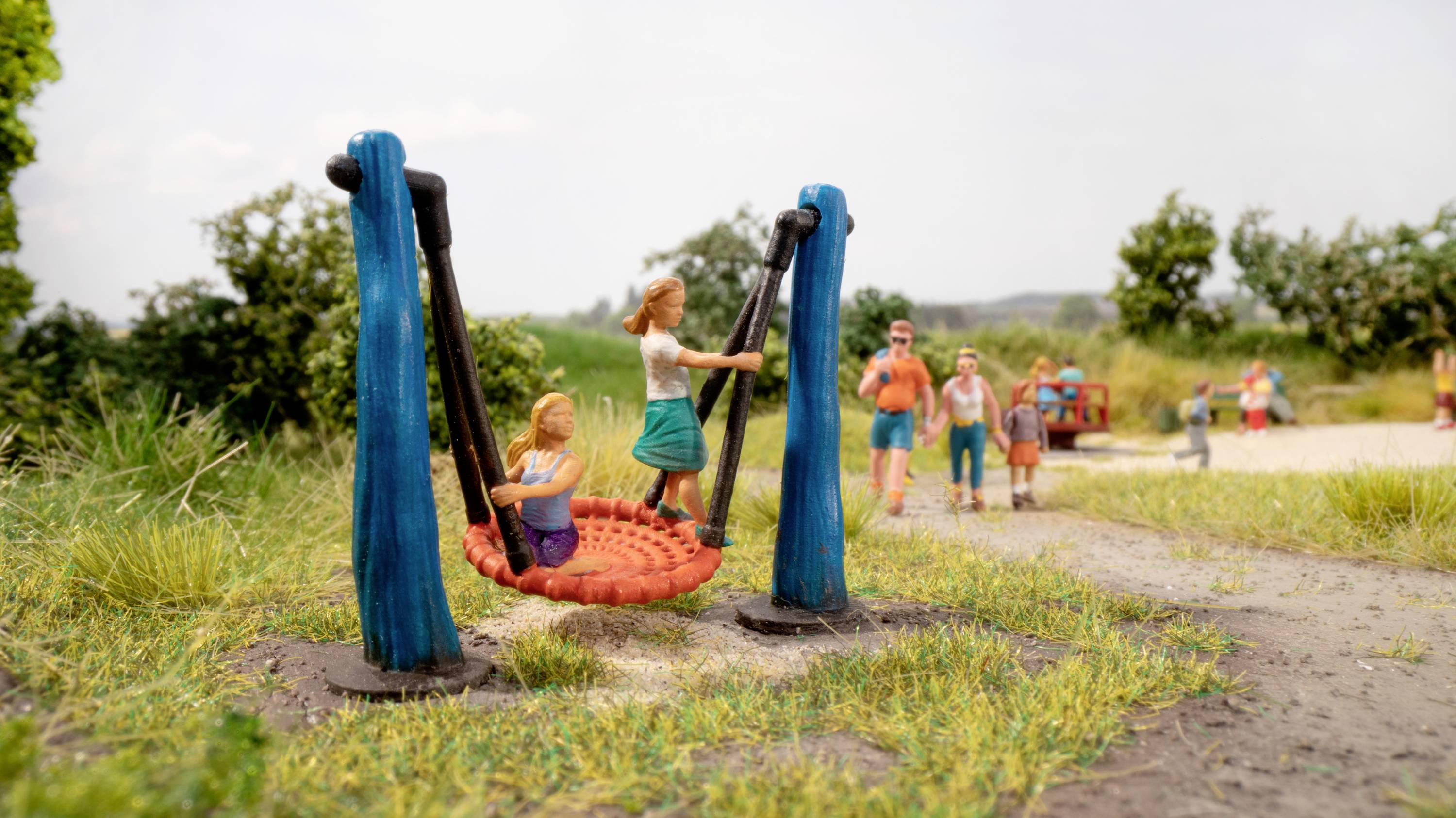 Spielplatzszene mit Kind und Erwachsenem auf Schaukel. Im Hintergrund Spaziergänger auf Weg und grüne Landschaft.