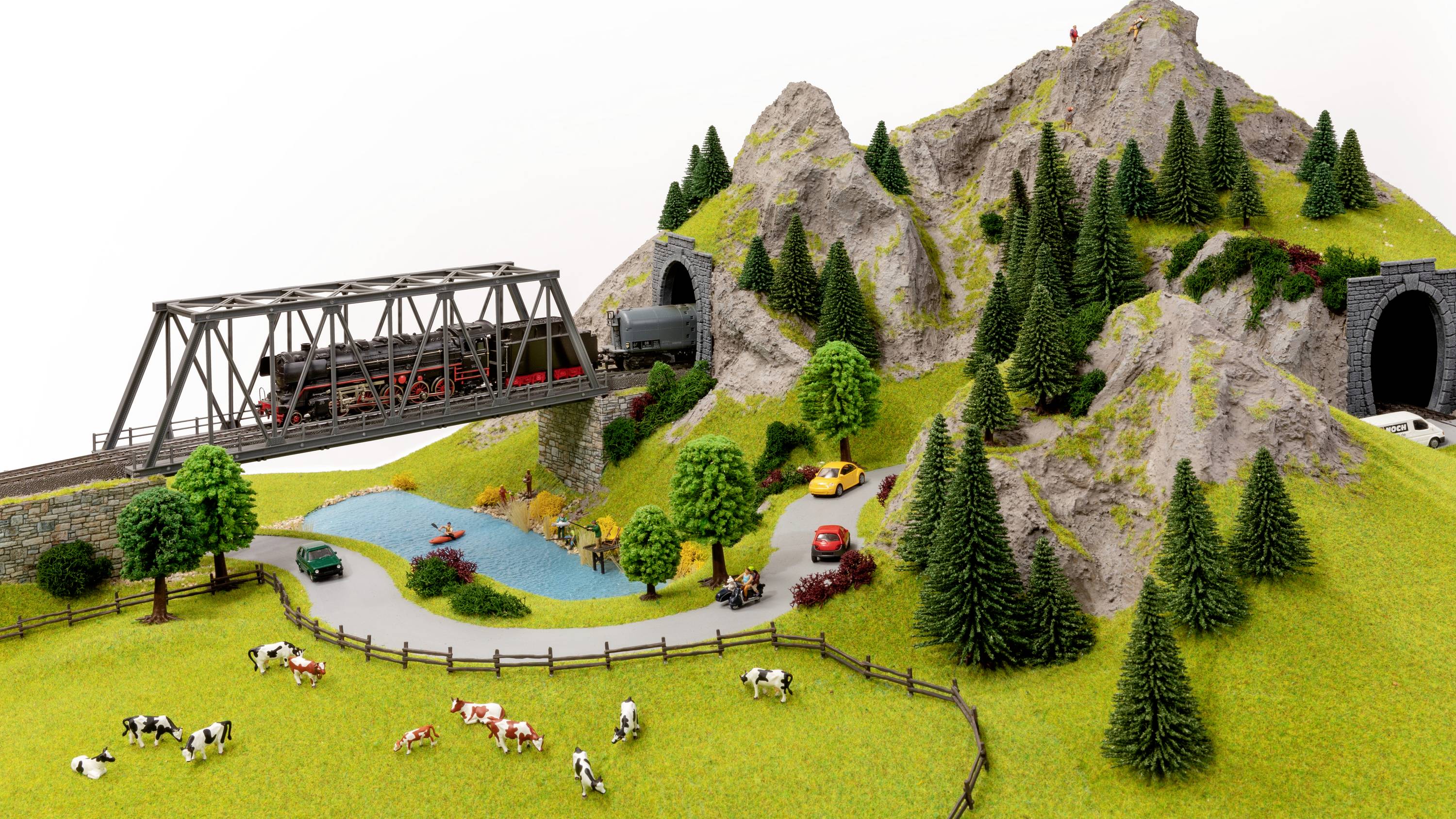 Eine Modelleisenbahn fährt über eine Brücke durch eine gebirgige Landschaft mit Tunneln. Kühe grasen auf einer Wiese neben einem See.