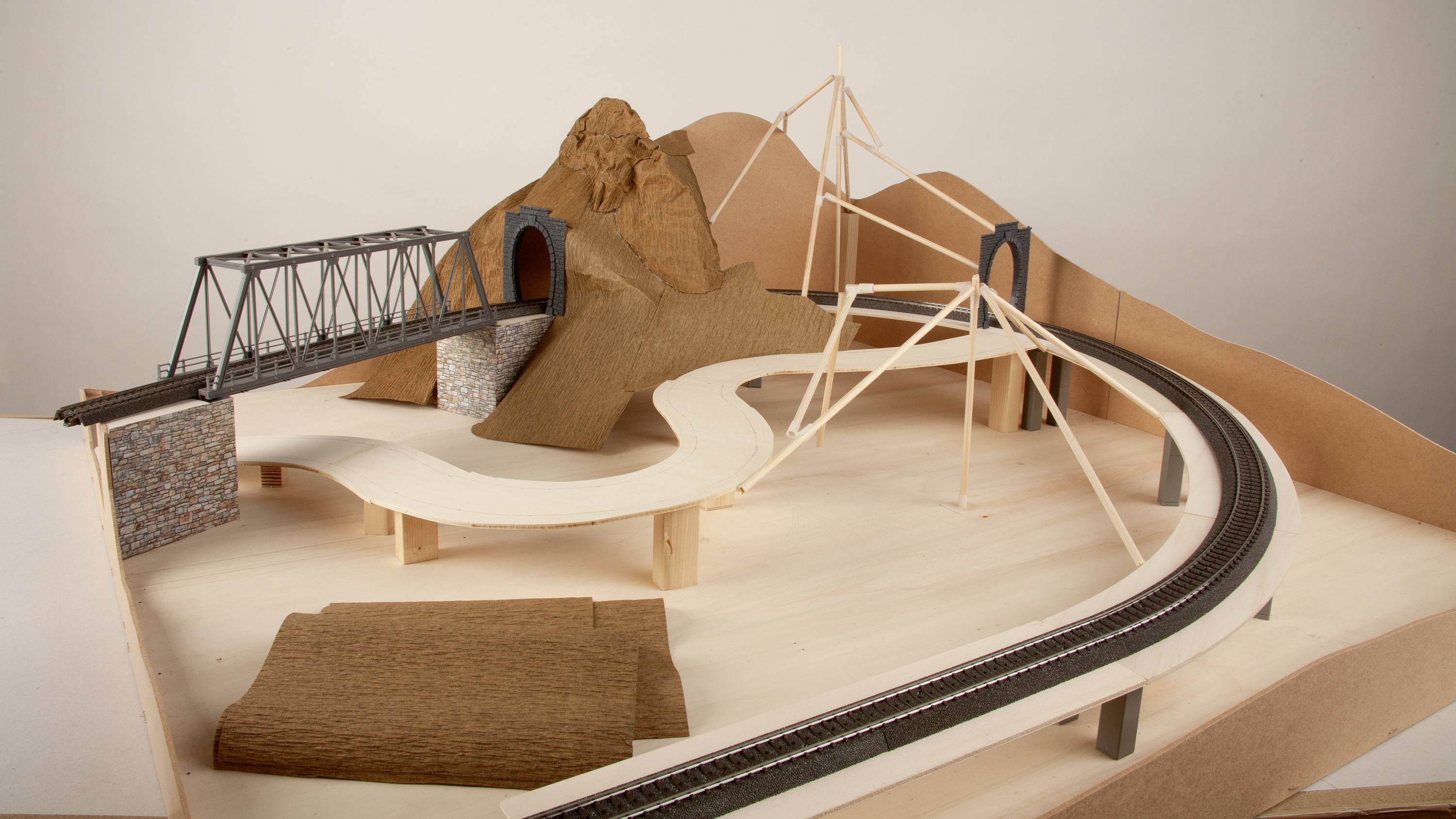 Modell einer Miniatureisenbahnlandschaft mit Brücken, einem Tunnel und einem Berg aus Karton und Holz. Schienen verlaufen über die Struktur.