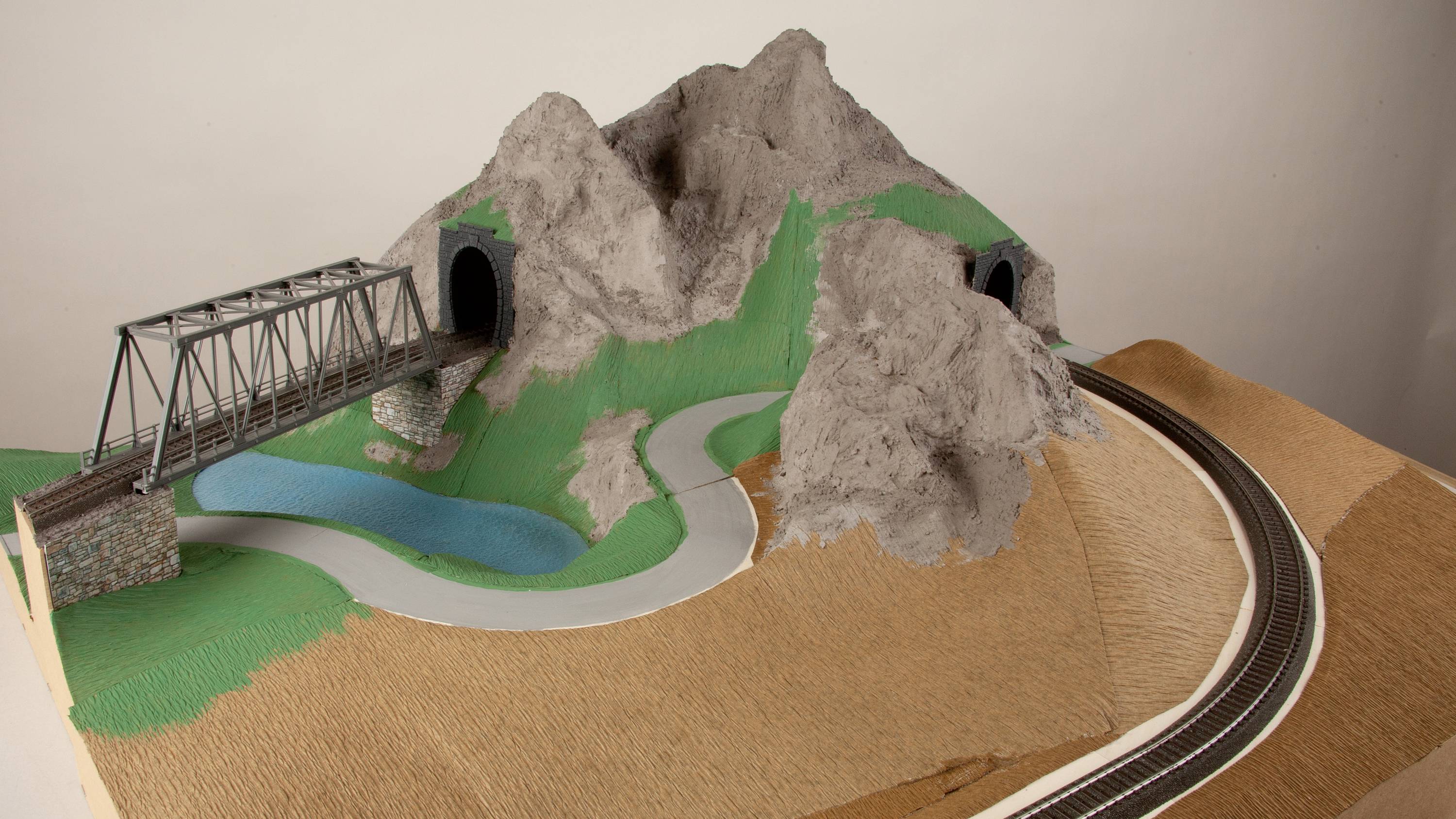Ein Modell einer Eisenbahnlandschaft mit Brücke und zwei Tunnel in einer Berglandschaft, umgeben von einer Straße und einem Teich.