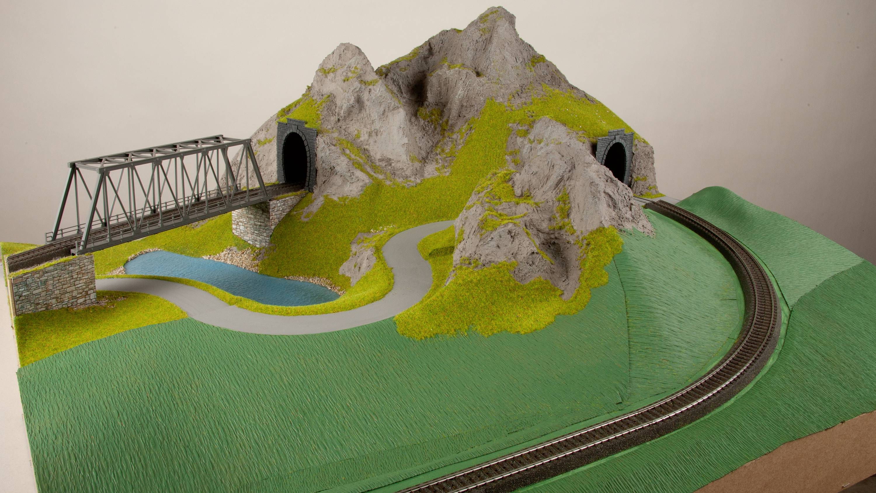 Modelleisenbahn-Landschaft mit einem Tunnel, einer Brücke, einer Straße und Schienen, die um einen Hügel führen.