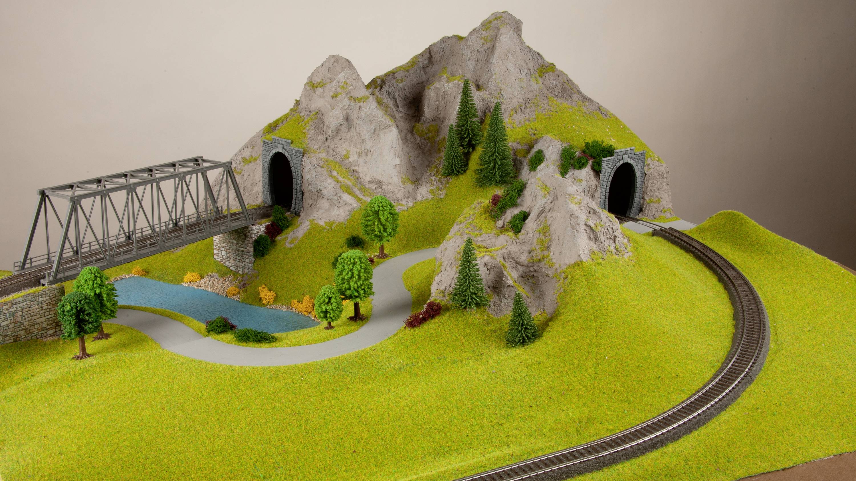 Modelleisenbahnlandschaft mit Brücke, zwei Tunnel in Hügeln, Wald, Wiesen und Bahngleisen; zeigt realistische Miniaturszene.