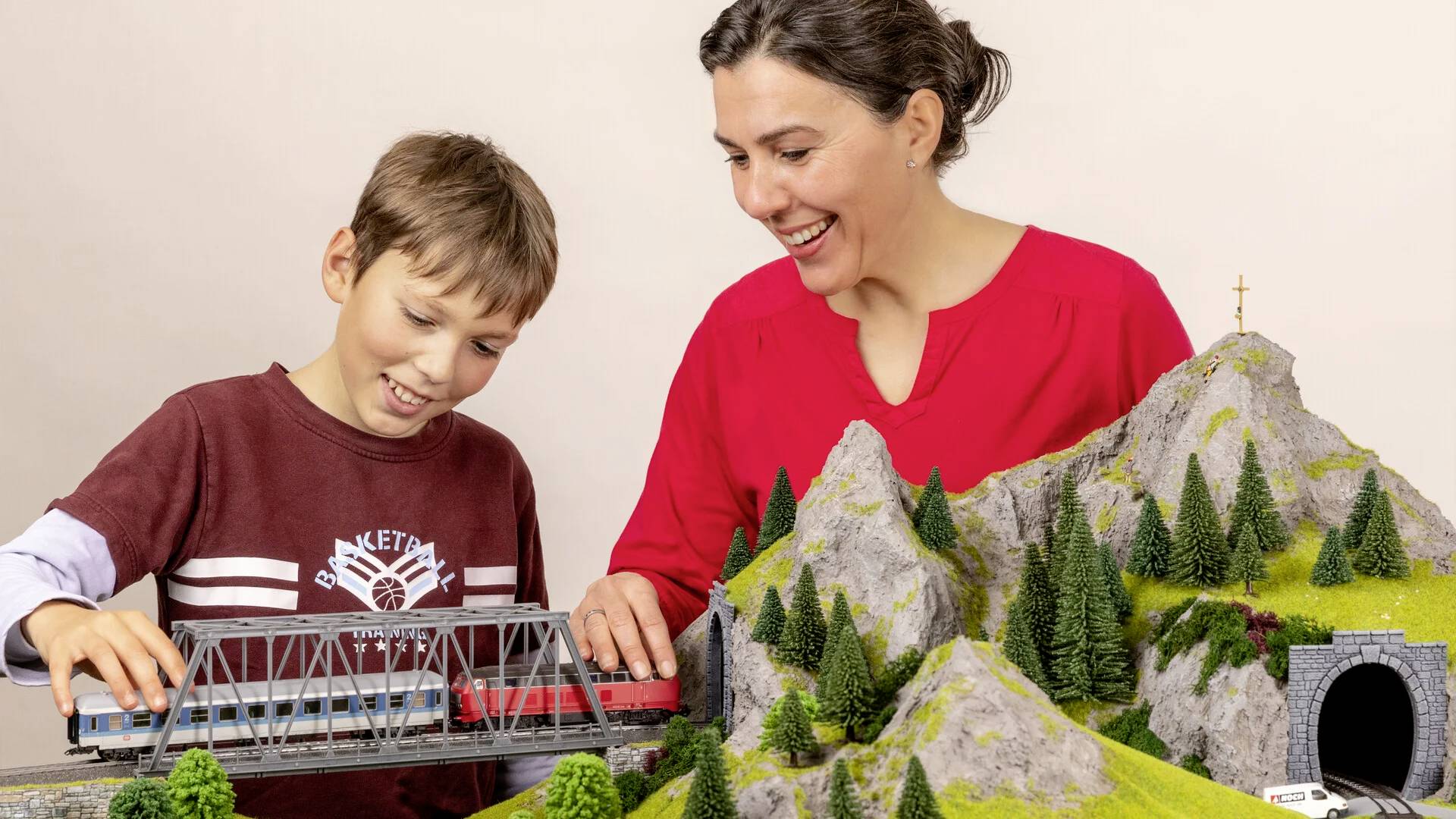 Eine Frau und ein Junge spielen gemeinsam mit einer Modelleisenbahn auf einer Landschaft mit Bergen, einer Brücke und einem Tunnel.
