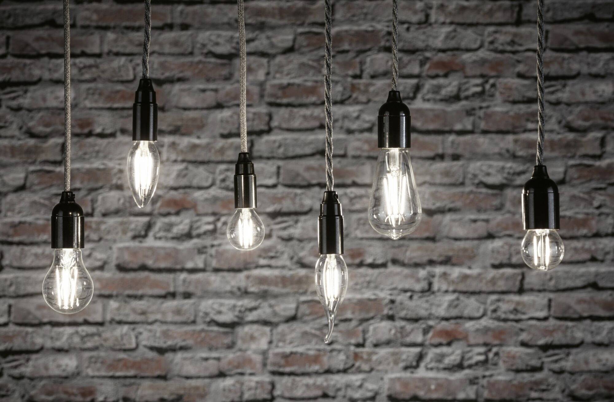 Mehrere hängende Glühbirnen leuchten vor einer Wand aus rotem Backstein und erzeugen eine industrielle Atmosphäre.