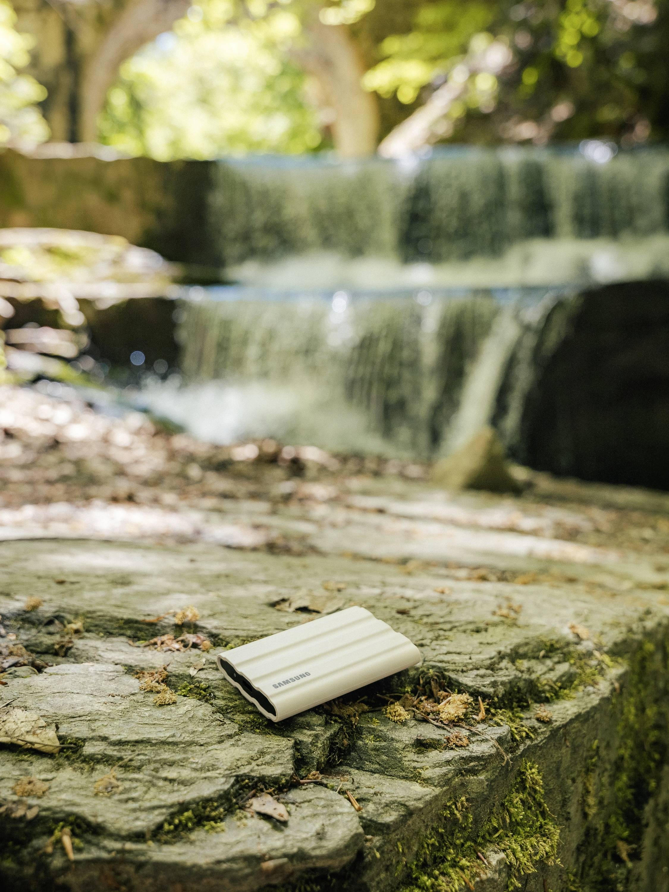 Ein kleines weißes Notizbuch liegt auf einem Felsen im Vordergrund, im Hintergrund ist ein natürlicher Wasserfall im Wald zu sehen.