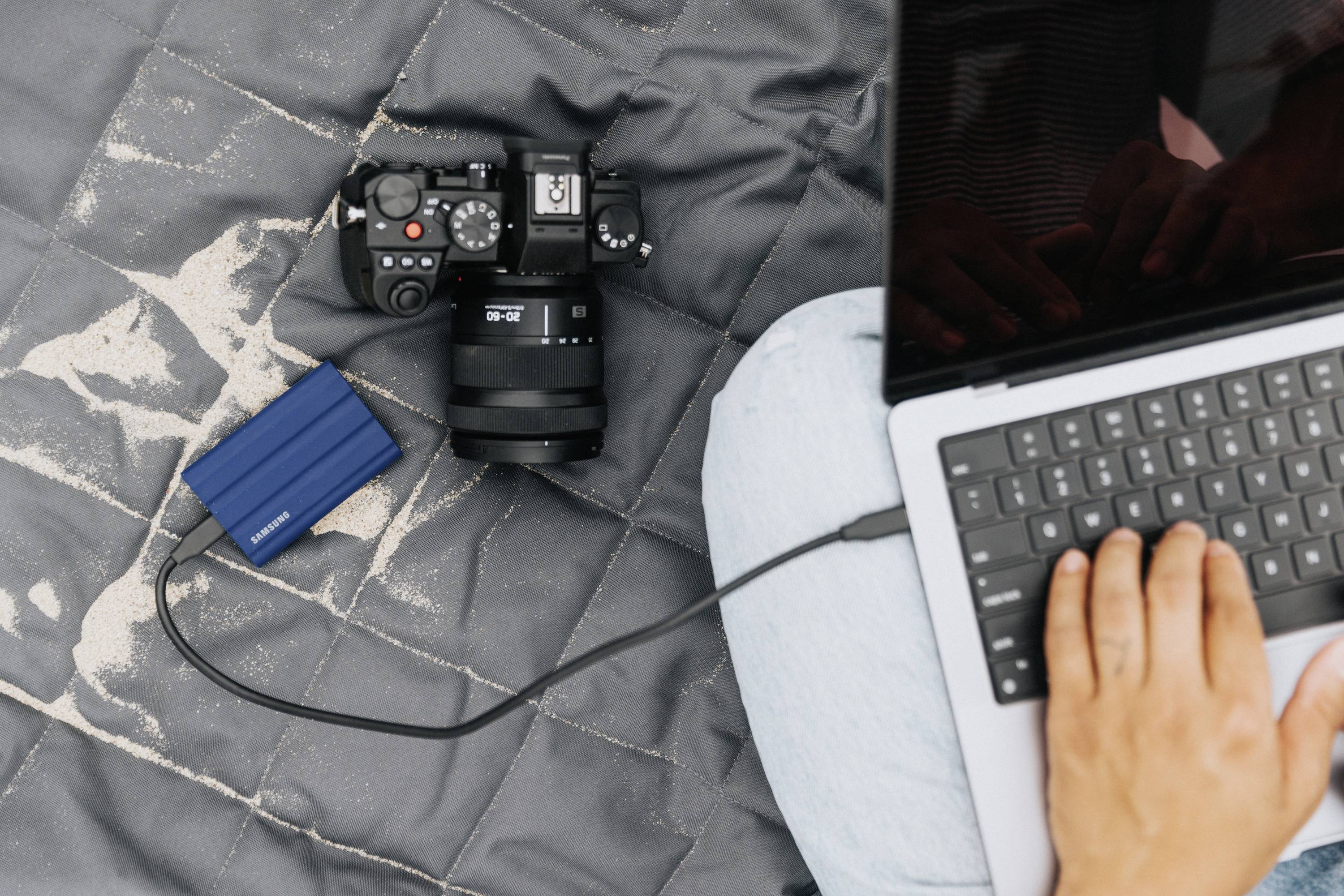 Eine Person arbeitet an einem Laptop, der an eine externe Festplatte angeschlossen ist. Daneben liegt eine Kamera auf einem grauen Tuch.