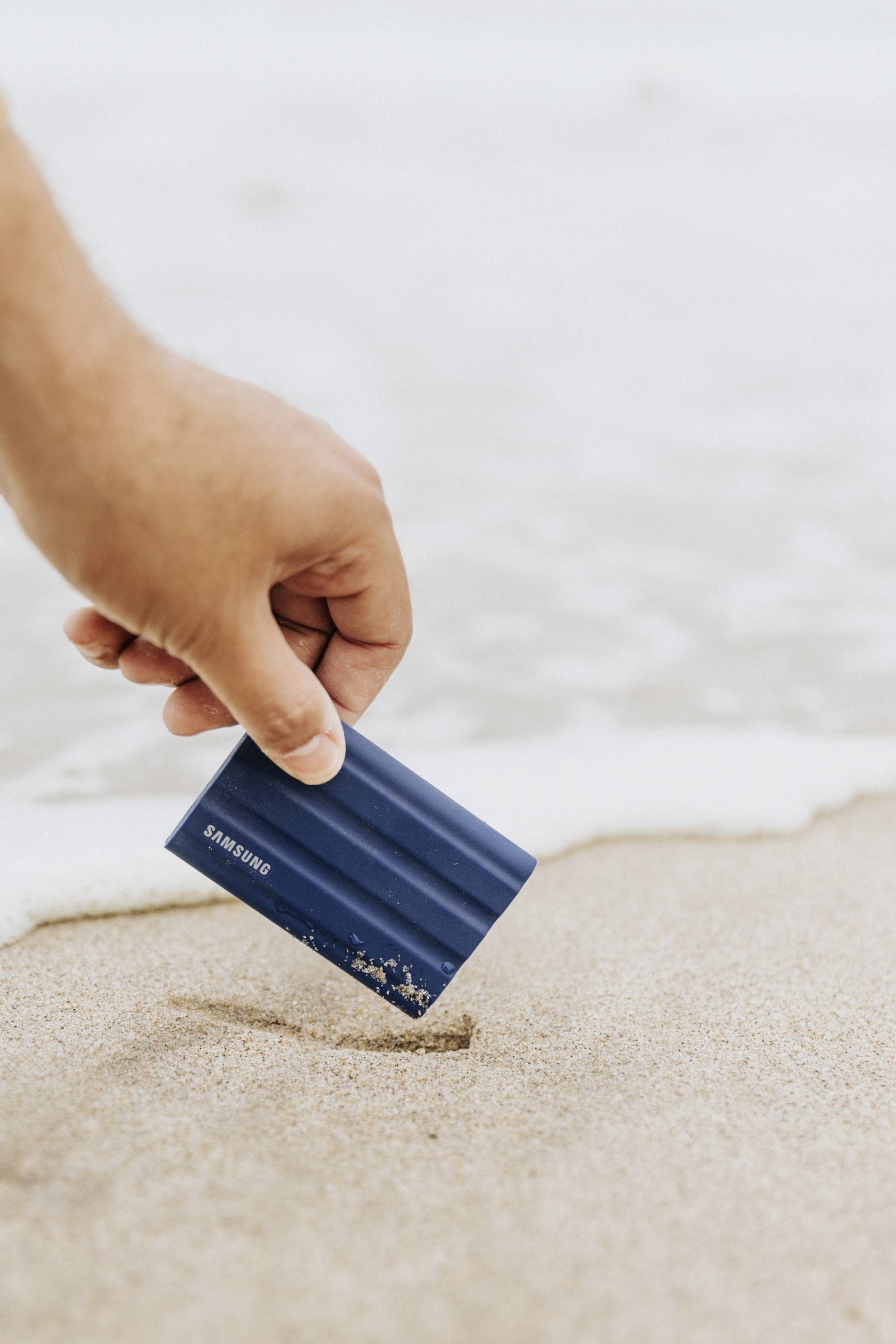 Eine Hand hebt ein externes Festplattenlaufwerk vom Sandstrand auf, im Hintergrund ist Wasser zu sehen.