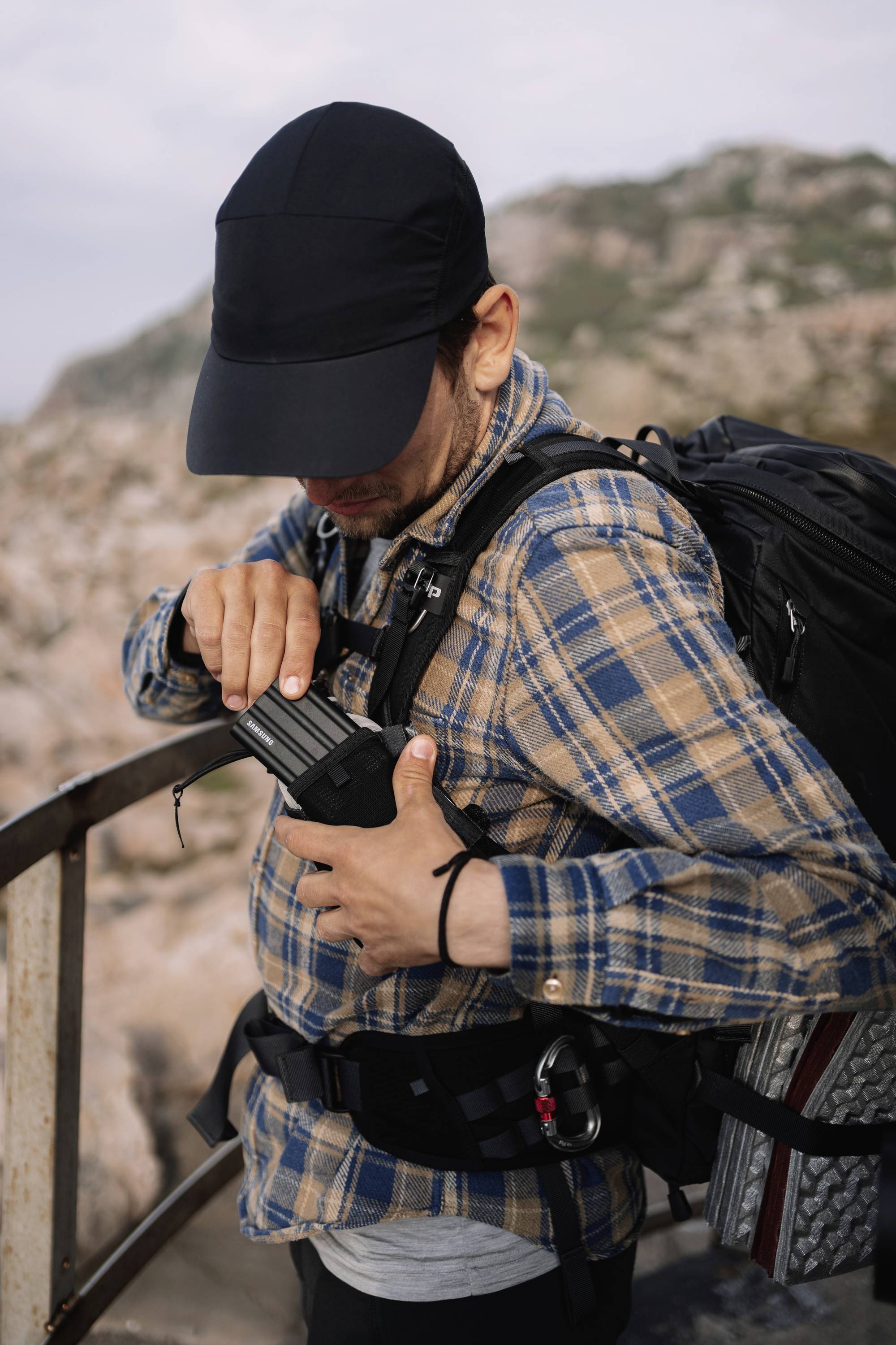 Eine Person steht auf einem Geländer in bergiger Landschaft, trägt karierte Kleidung, einen Rucksack und hantiert mit einem Gerät.