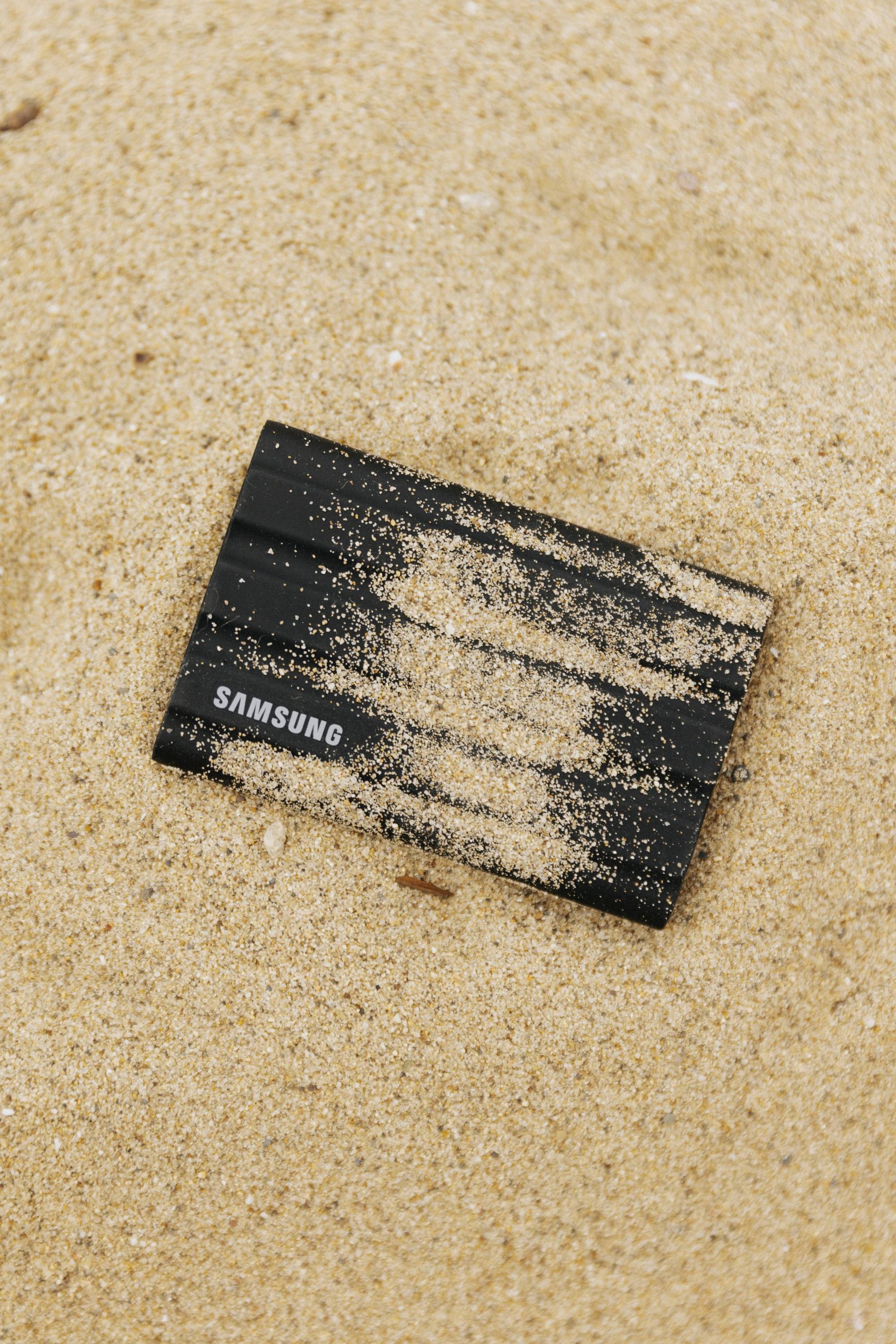 Eine schwarze Samsung-Festplatte liegt im Sand. Oben ist sie teilweise mit Sand bedeckt.