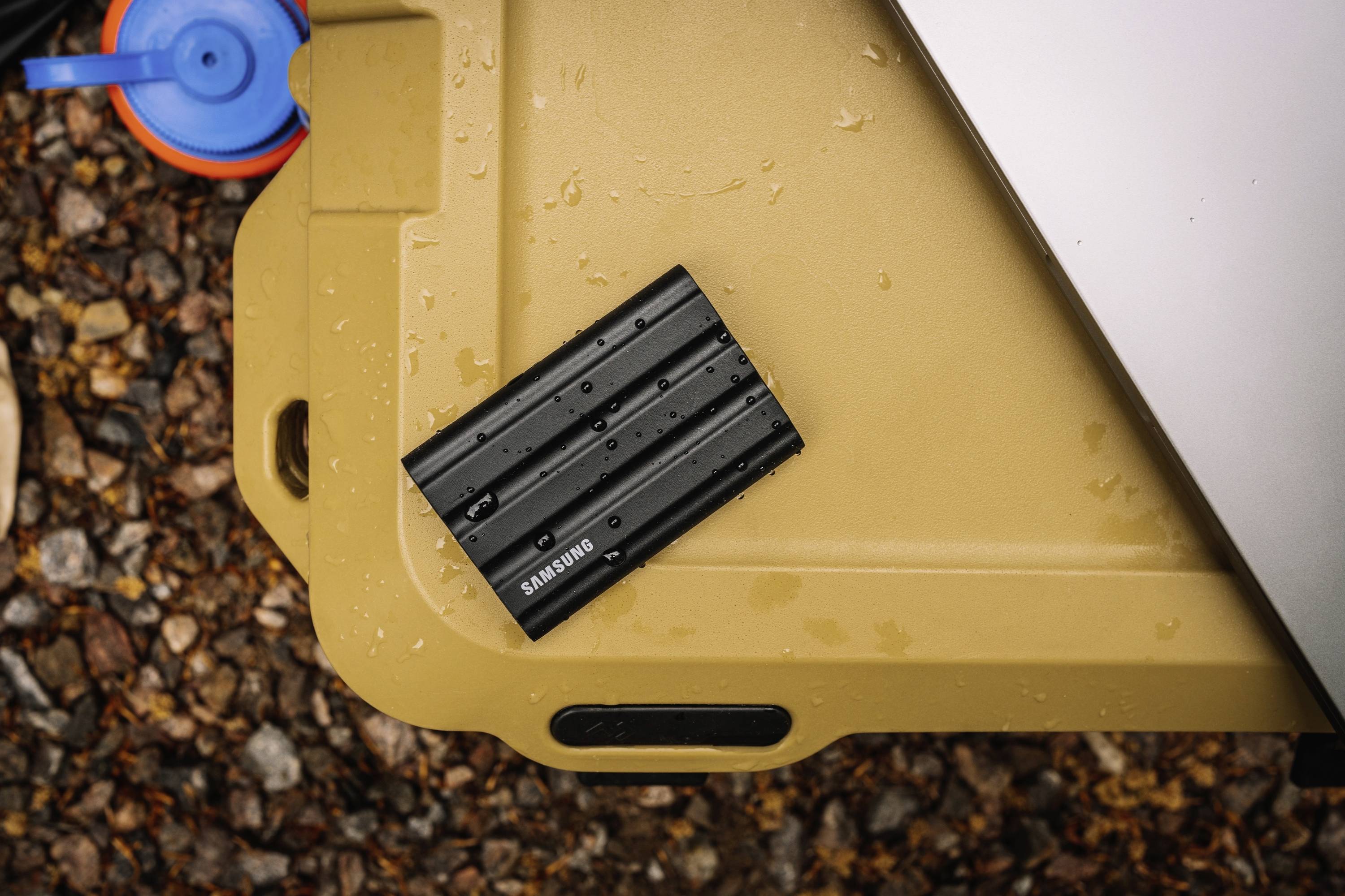 Externe Festplatte mit Wassertropfen darauf, platziert auf einer sandfarbenen Oberfläche neben einem blauen Deckel.