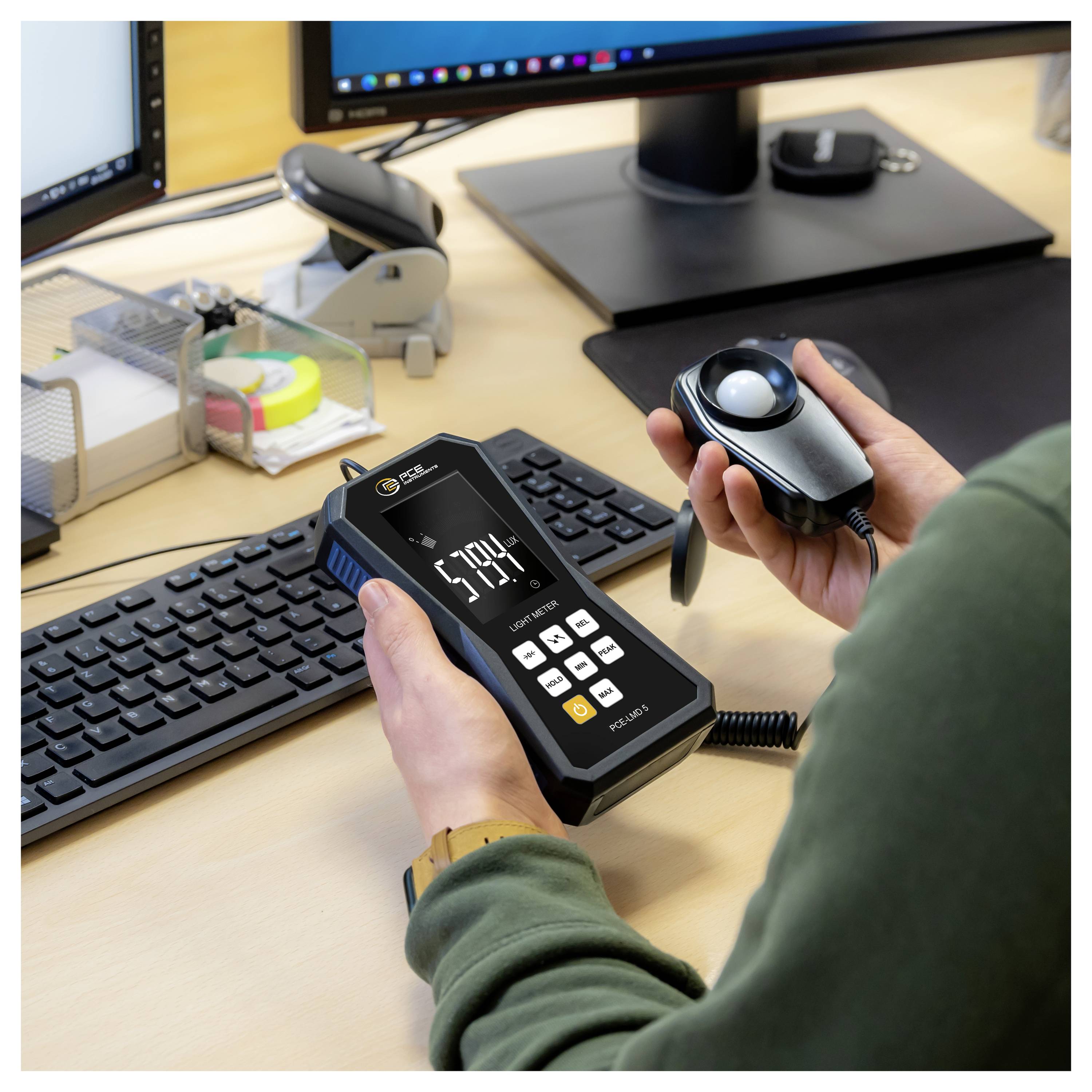 Person mit einem tragbaren Luxmeter und kabelgebundenem Sensor an einem Schreibtisch, der die Bildschirmhelligkeit in der Nähe eines Computermonitors und einer Tastatur misst.