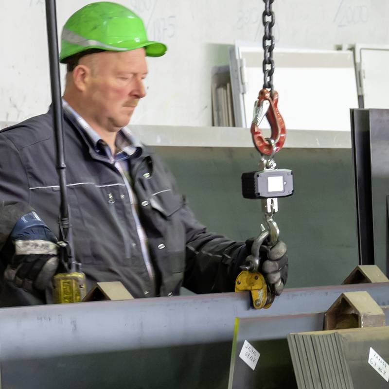 Ein Arbeiter mit grünem Helm bedient einen Kran in einer Industriehalle. Metallplatten werden bewegt. Sicherheitsschuhe und Handschuhe sichtbar.