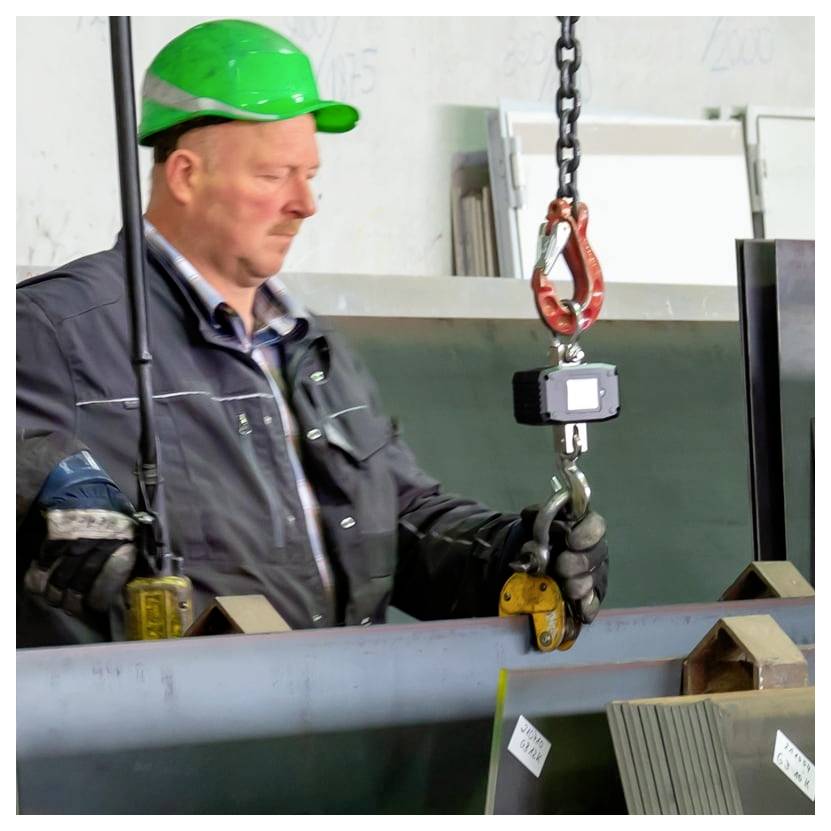 Fabrikarbeiter in einem grünen Schutzhelm benutzt einen Krahaken und eine Hebeklammer, um einen großen Metallbalken zu bewegen.